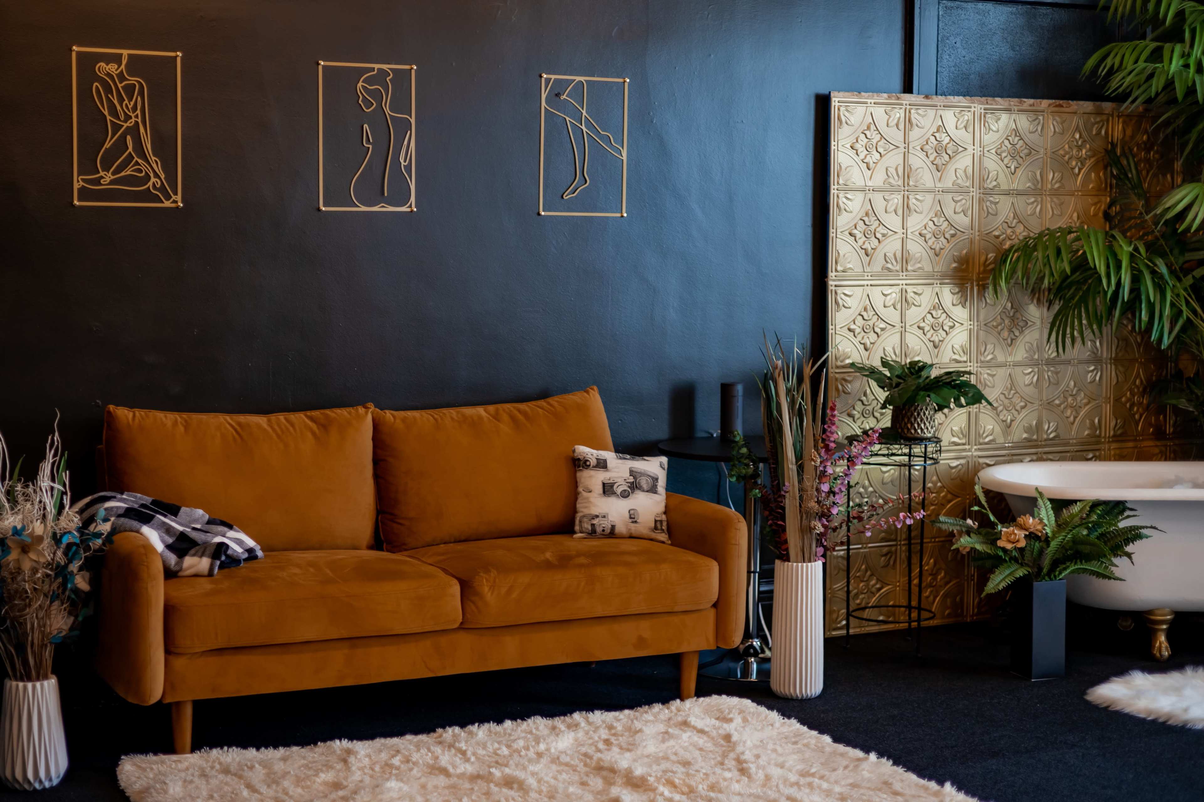 A cozy living area featuring a brown suede sofa, gold wall art, a small white rug, and a bathtub surrounded by plants.