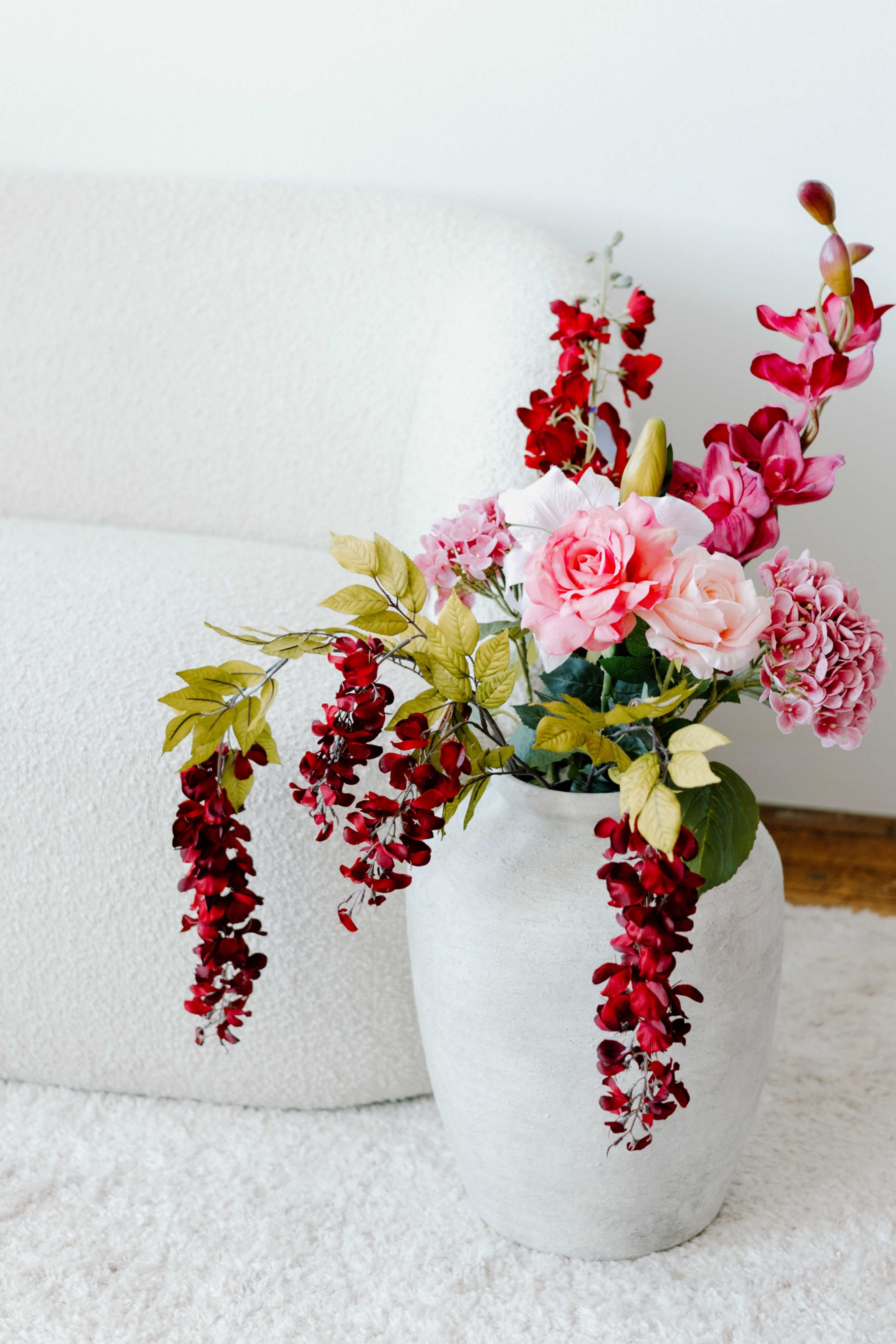 A concrete vase holds a colorful arrangement of artificial flowers, including pink roses and red blooms, beside a textured white sofa on a light rug.