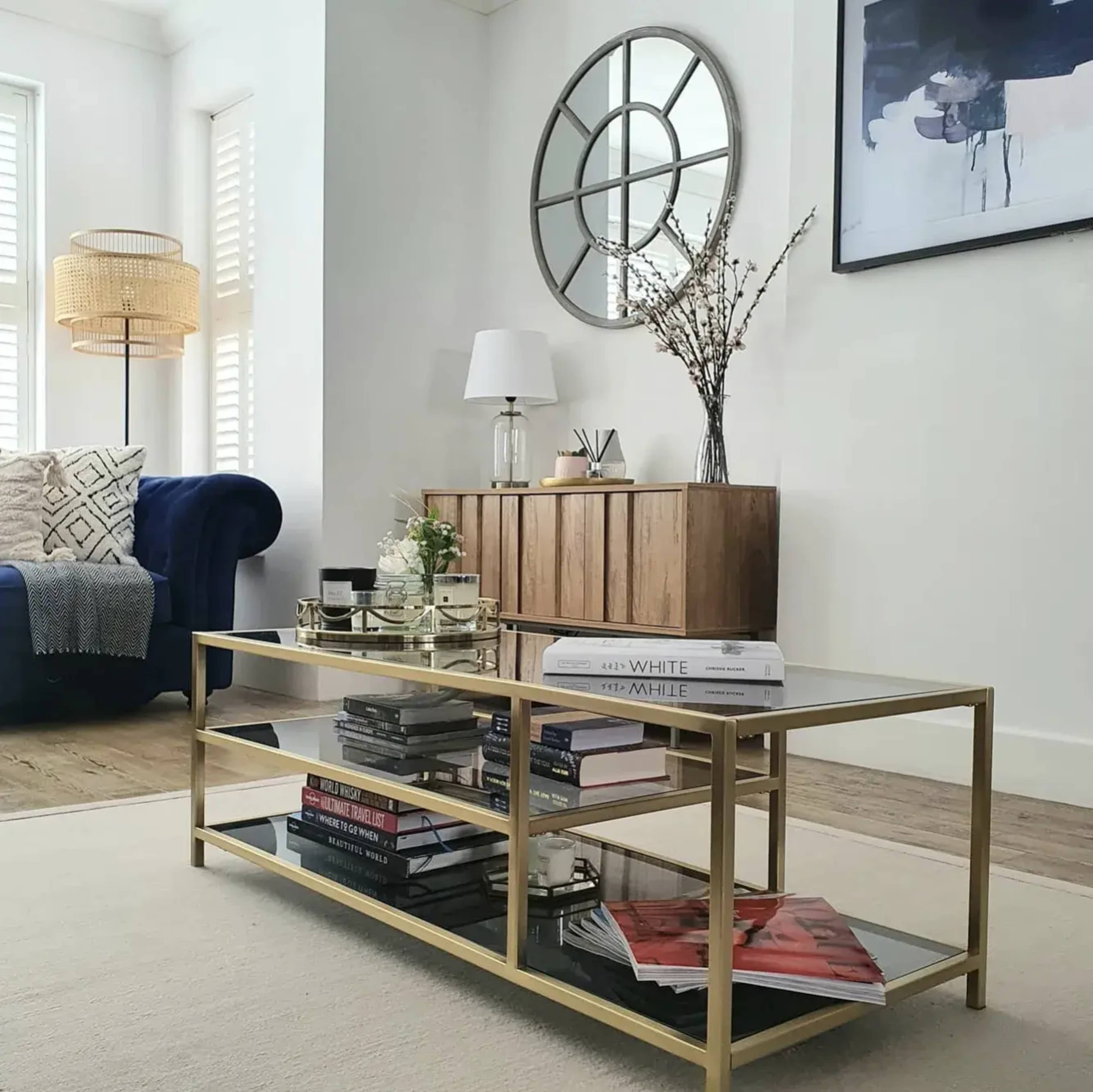 A living room featuring a glass-top coffee table with books and decor, a blue sofa, a round mirror on the wall, and a wooden cabinet.