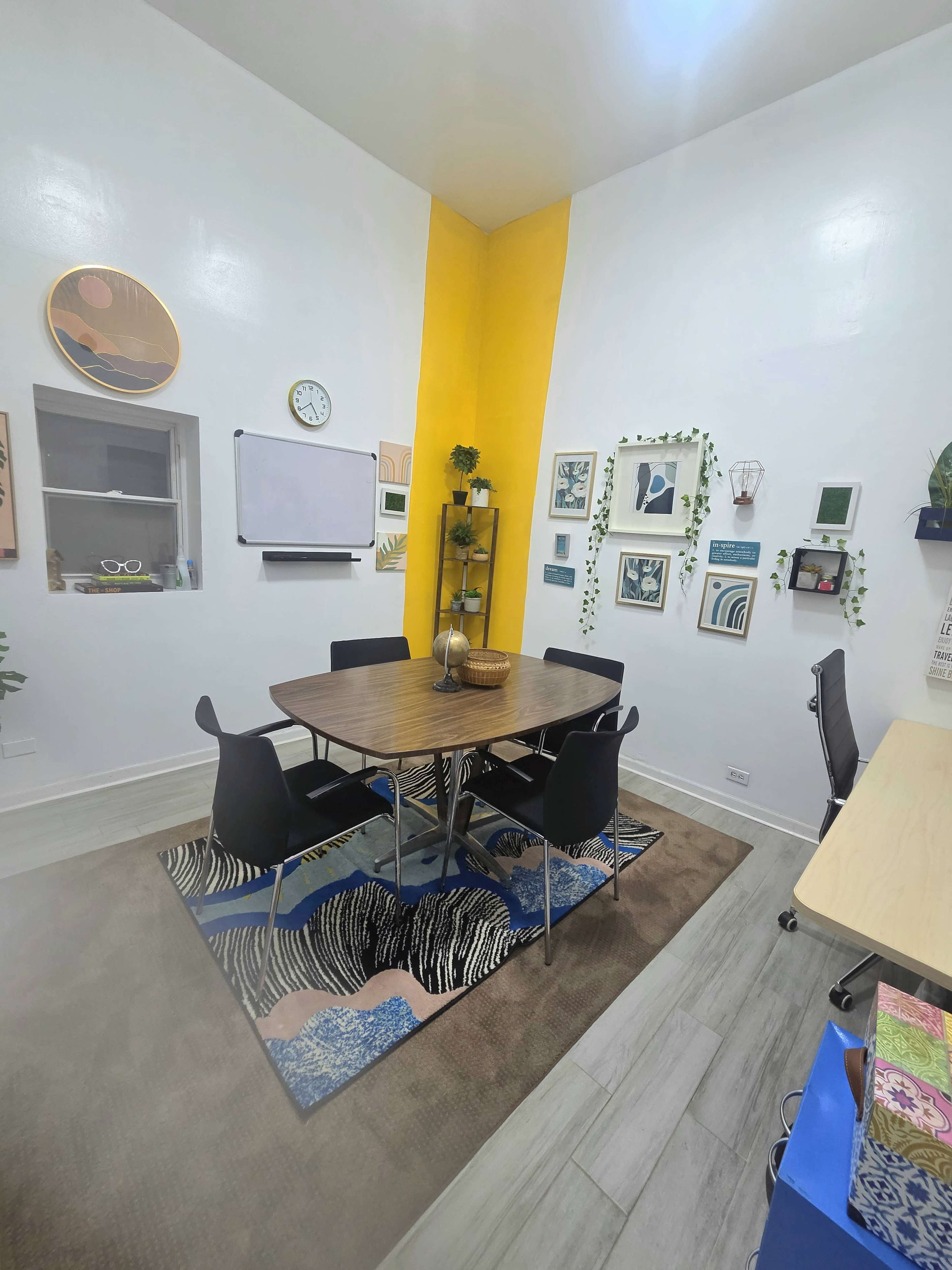 The image shows a bright, modern meeting room with a wooden table surrounded by black chairs, a large yellow accent wall, and various framed artworks on the walls.