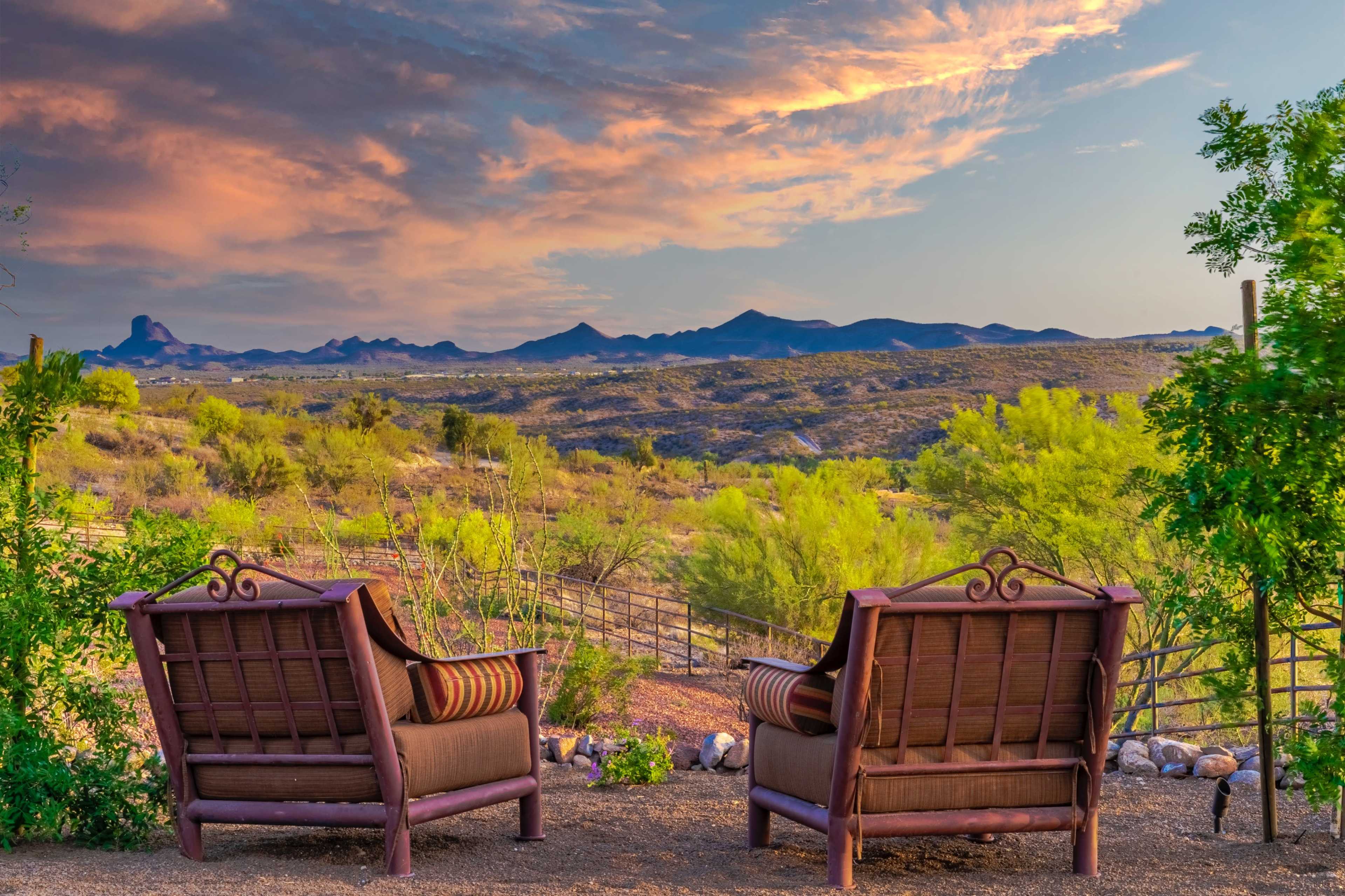 Two comfortable chairs face a scenic view of rolling hills and a colorful sunset sky in a desert landscape.
