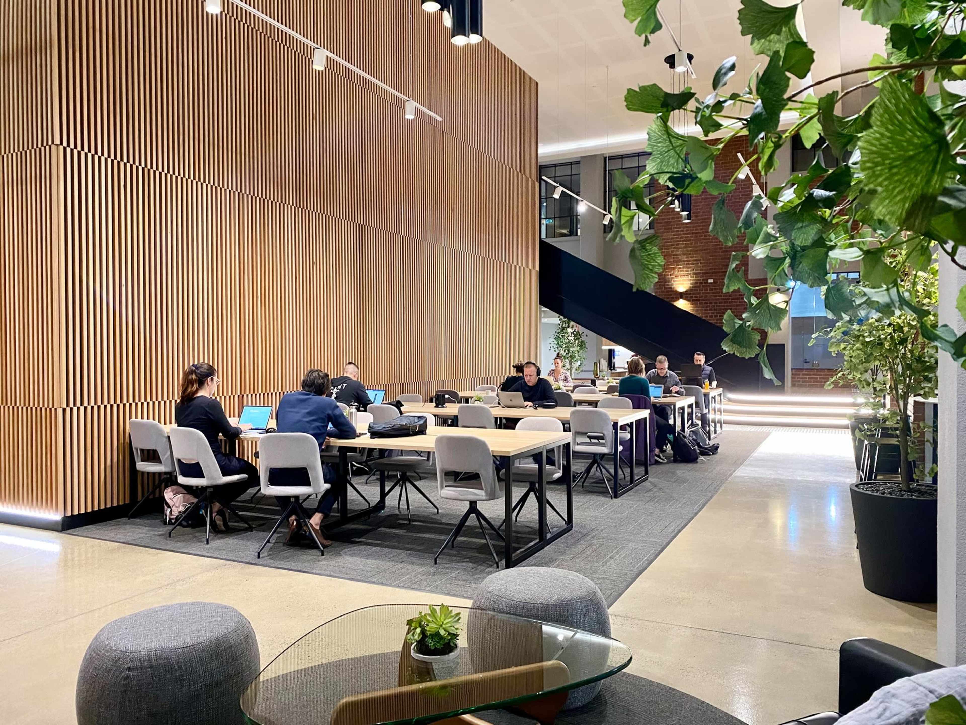 A modern workspace features several individuals sitting at tables with laptops, surrounded by wooden walls and plants.