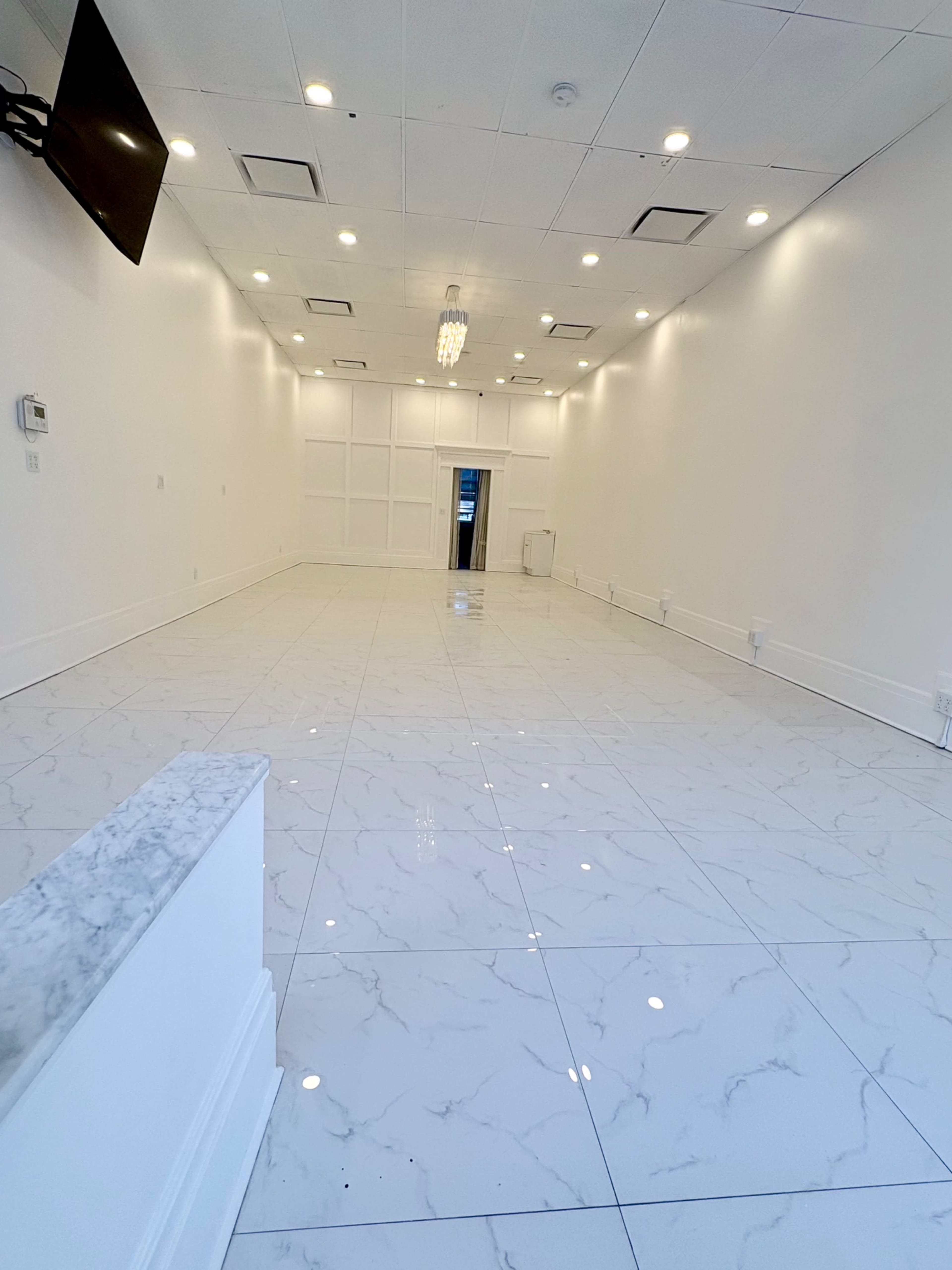 The image shows a spacious, empty room with glossy white marble flooring, white walls, and a chandelier hanging from the ceiling.