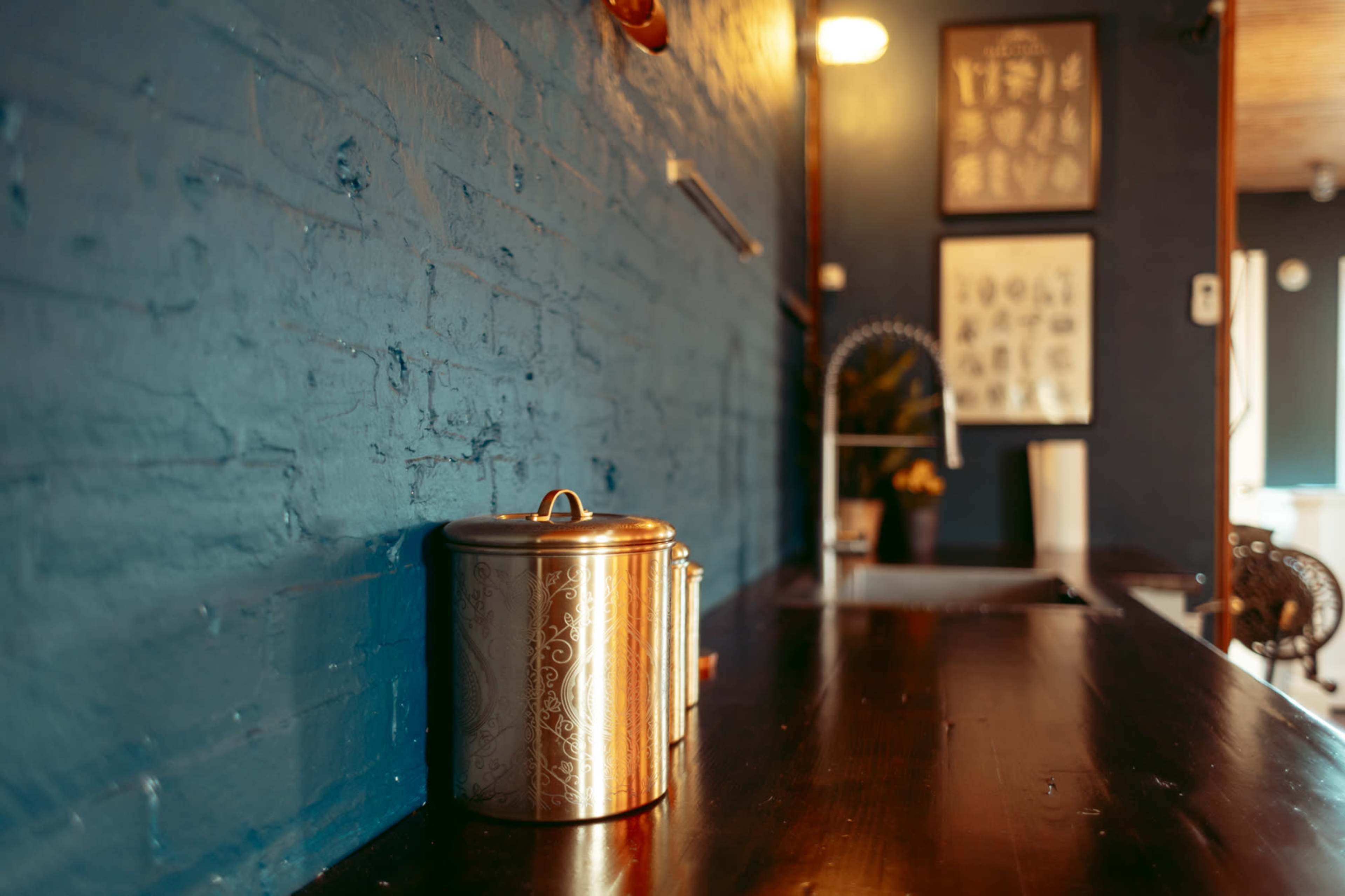 A copper canister sits on a wooden table against a blue brick wall, with framed artwork visible in the background.