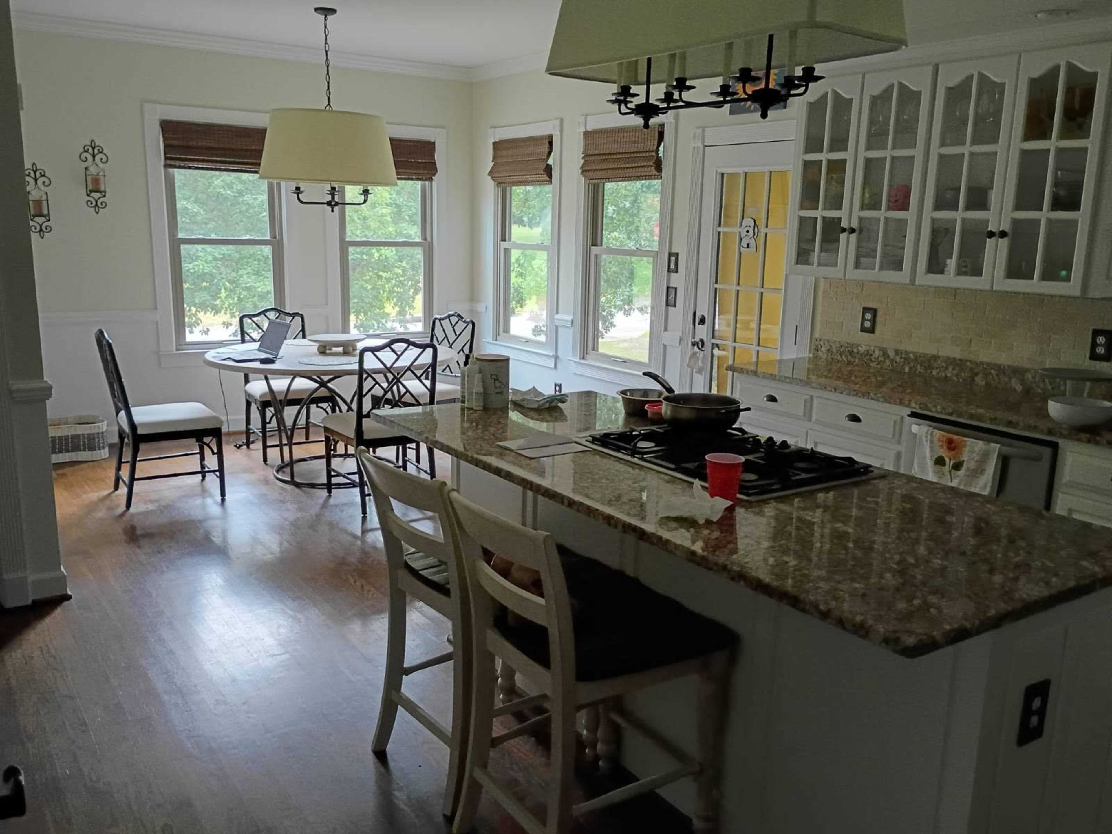 The image shows a spacious, well-lit kitchen featuring a central island with bar stools, a dining area with a round table, and large windows overlooking a green view.
