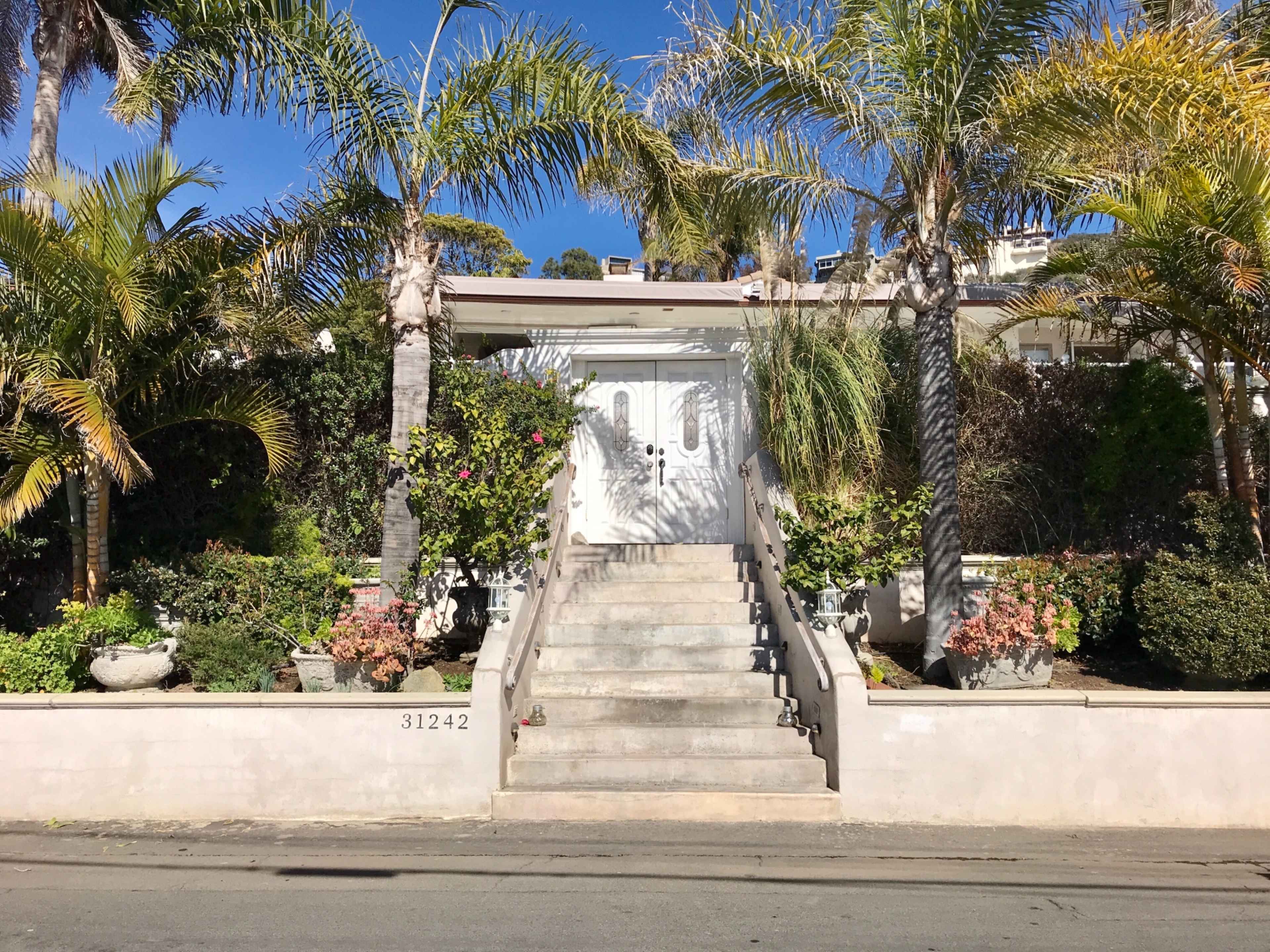 A white front door is framed by palm trees and lush greenery, with a wide staircase leading up to it.