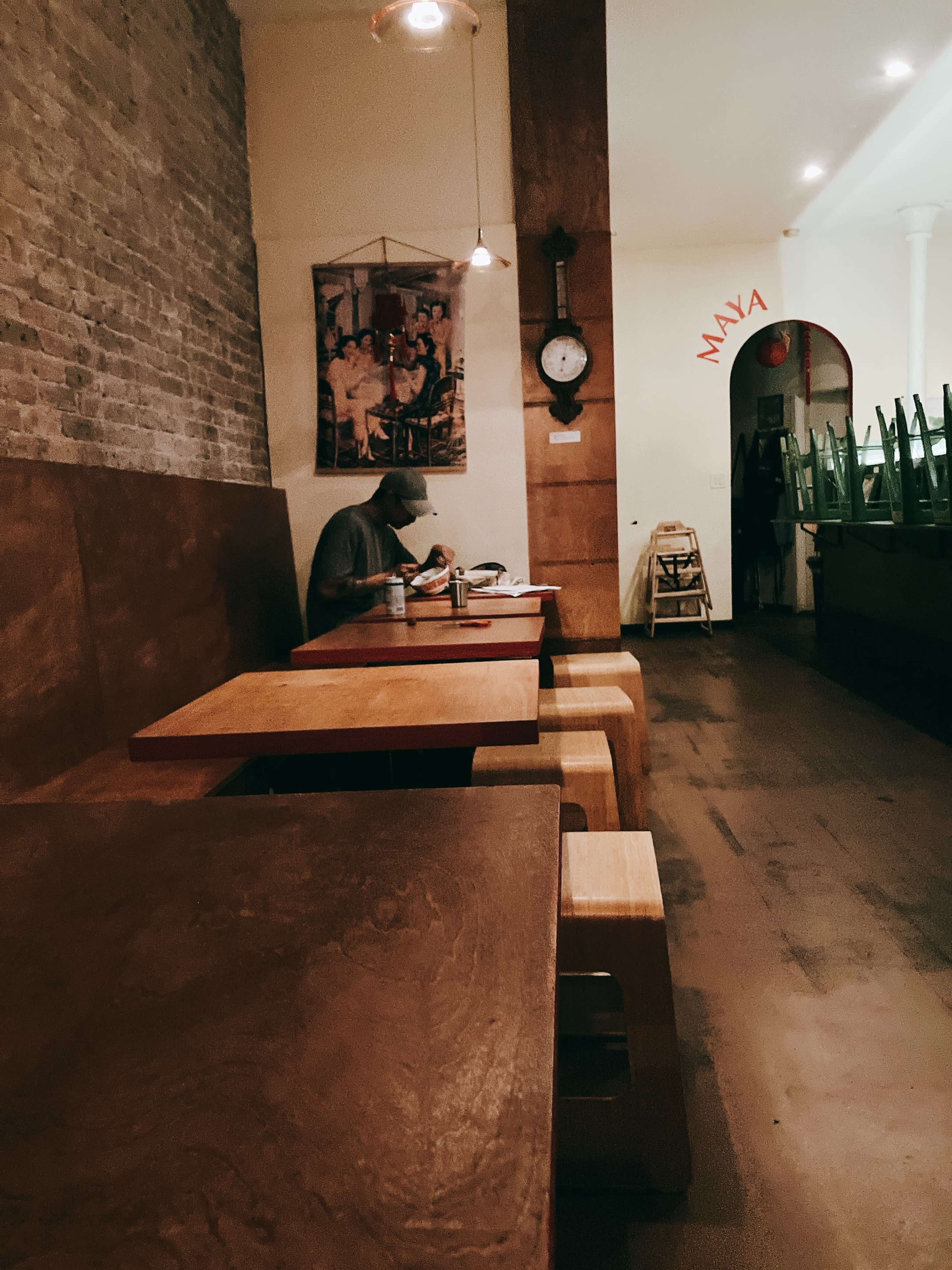 A solitary person sits at a wooden table in a sparsely furnished restaurant with exposed brick walls and minimal decor.