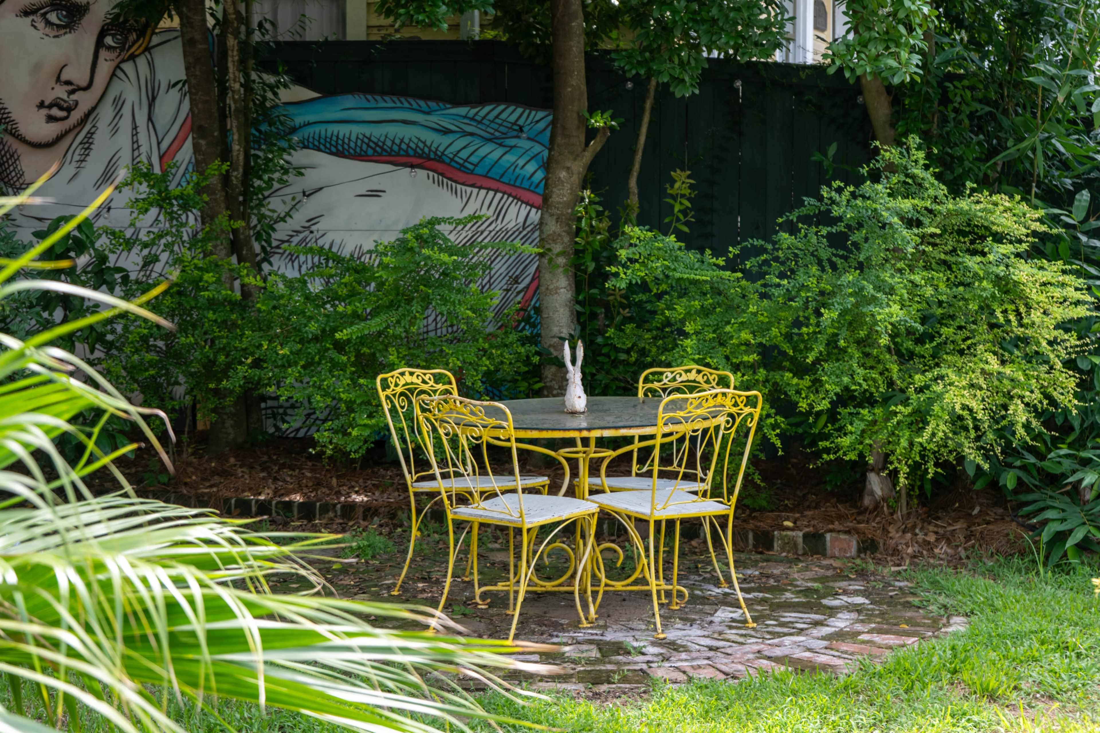 A small round table with four yellow chairs is located on a stone patio surrounded by lush greenery and a mural in the background.