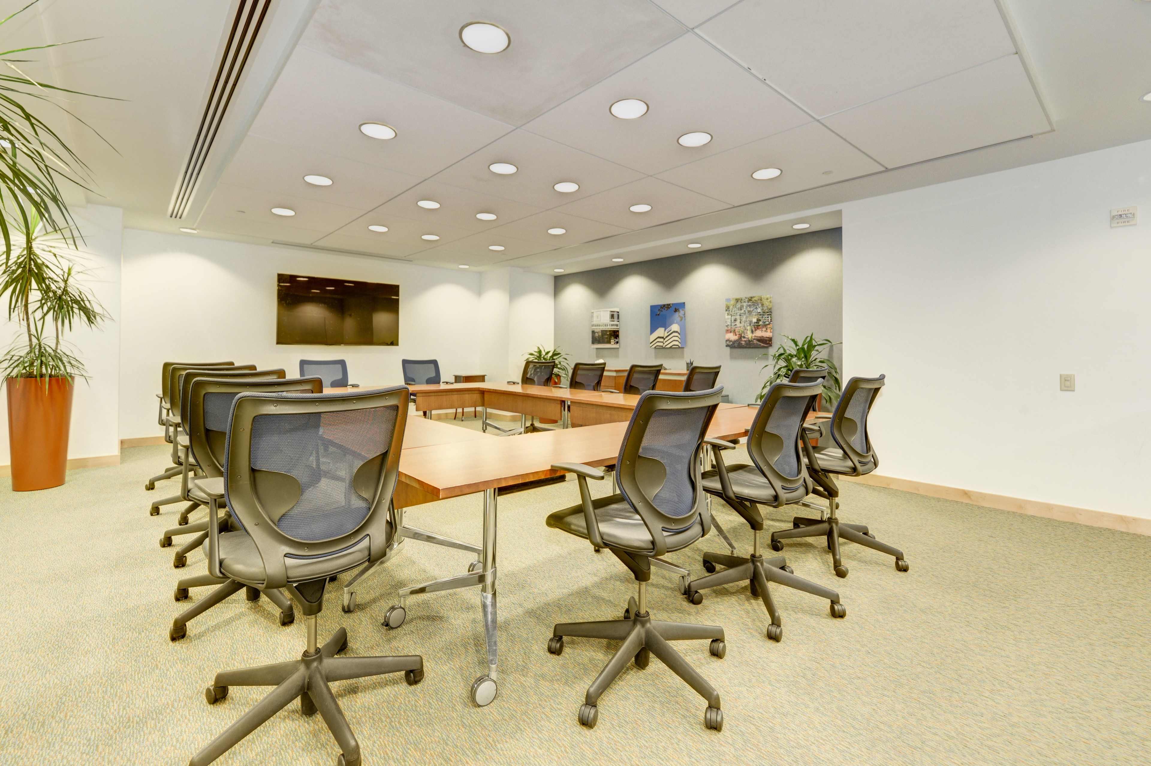 A spacious conference room features a large wooden table surrounded by black swivel chairs, with potted plants and a wall-mounted screen at one end.