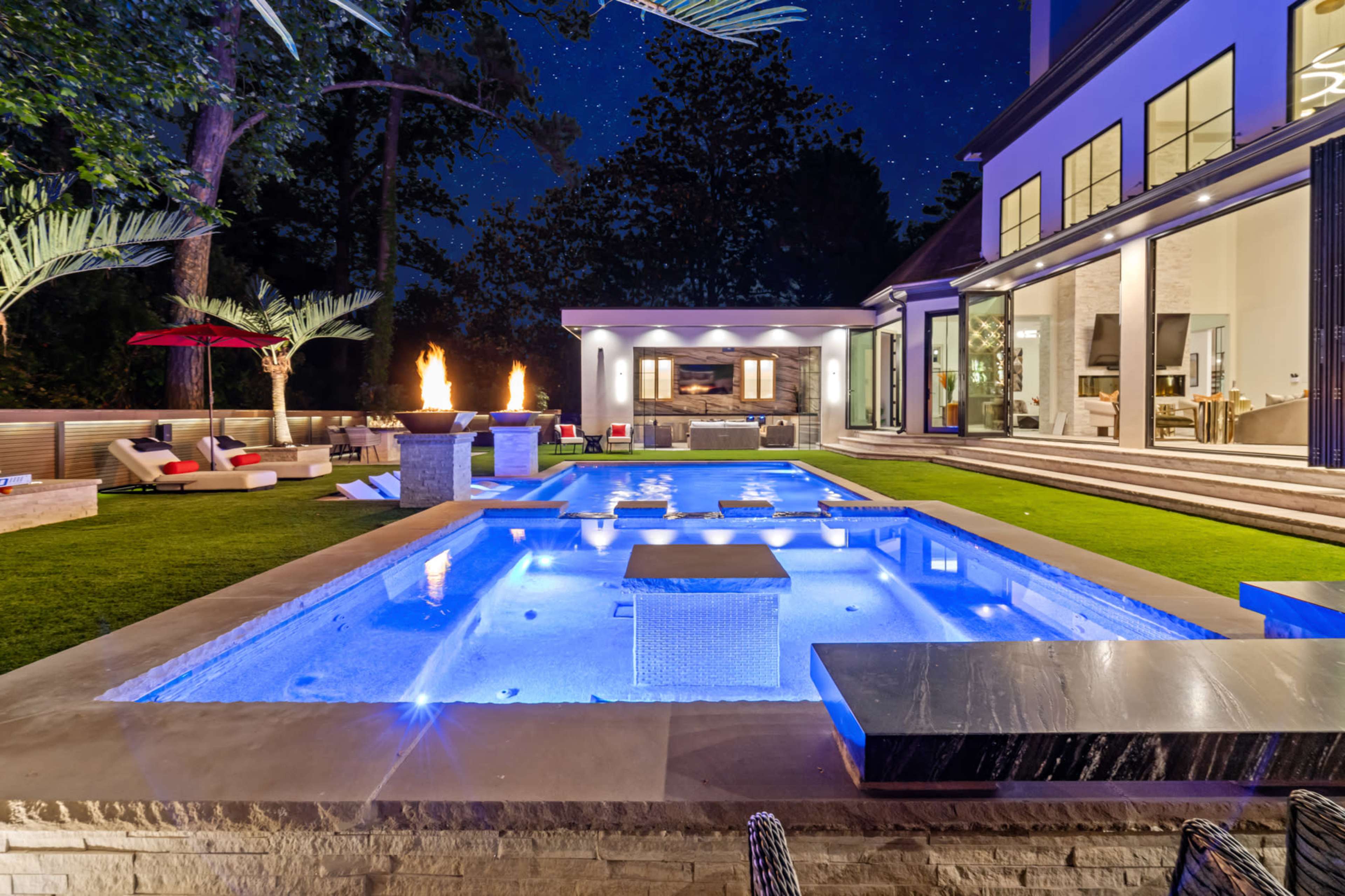 The image shows a luxurious backyard at night featuring a well-lit pool, modern outdoor furniture, and a fire pit surrounded by landscaped greenery.