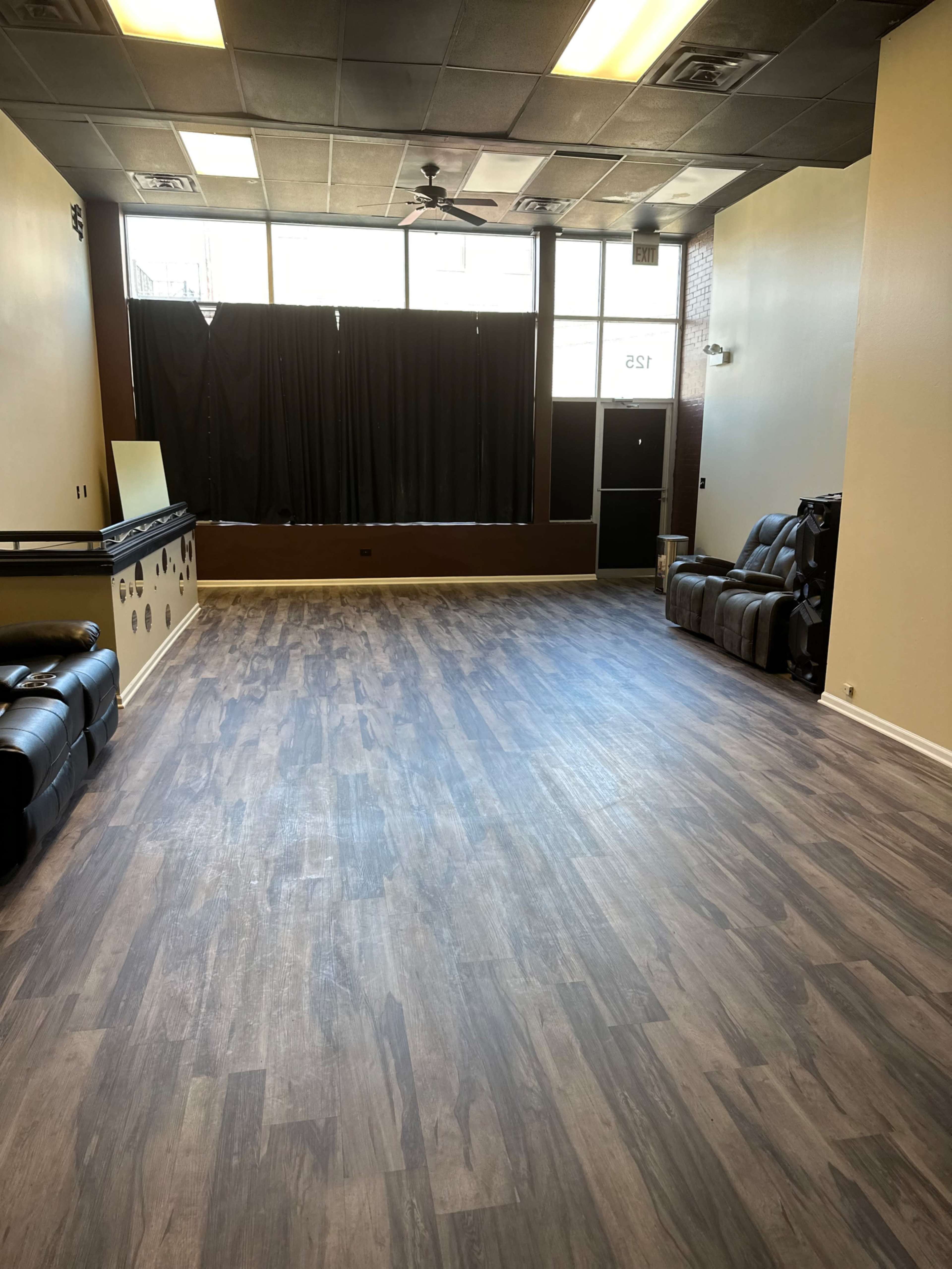 The image shows a spacious room with wooden laminate flooring, empty except for a few pieces of dark furniture along the walls and large windows covered by black curtains.