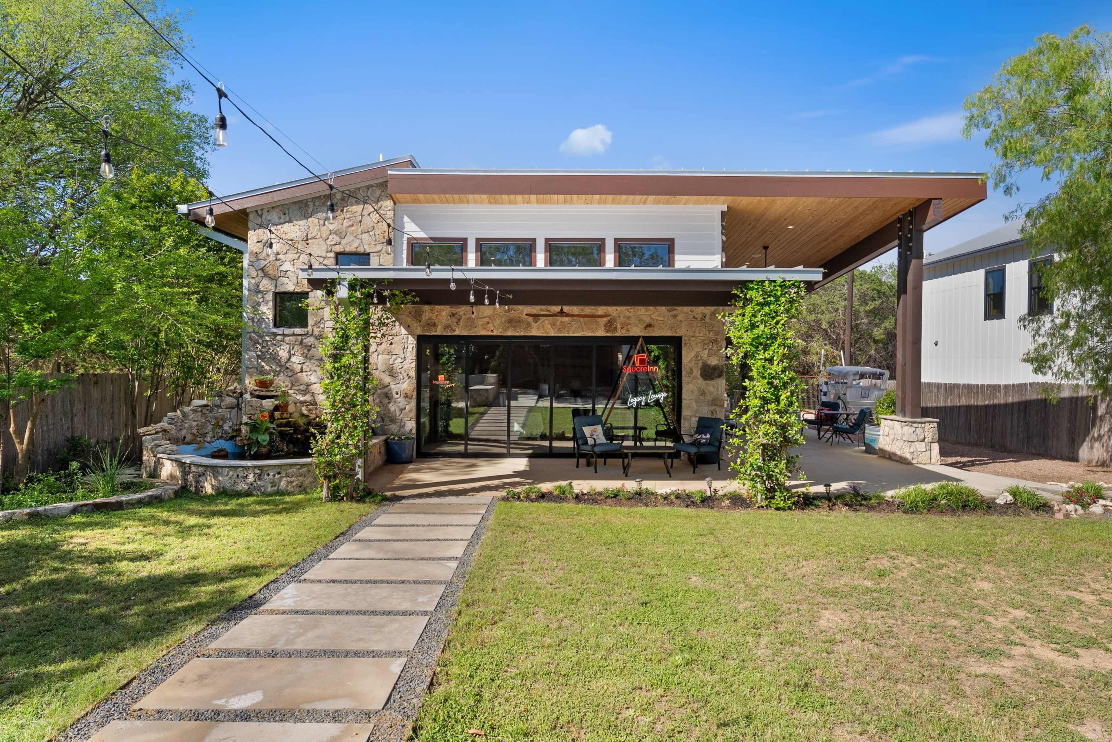 A modern house features a stone facade, large glass windows, and a covered porch with hanging lights, set in a landscaped yard.