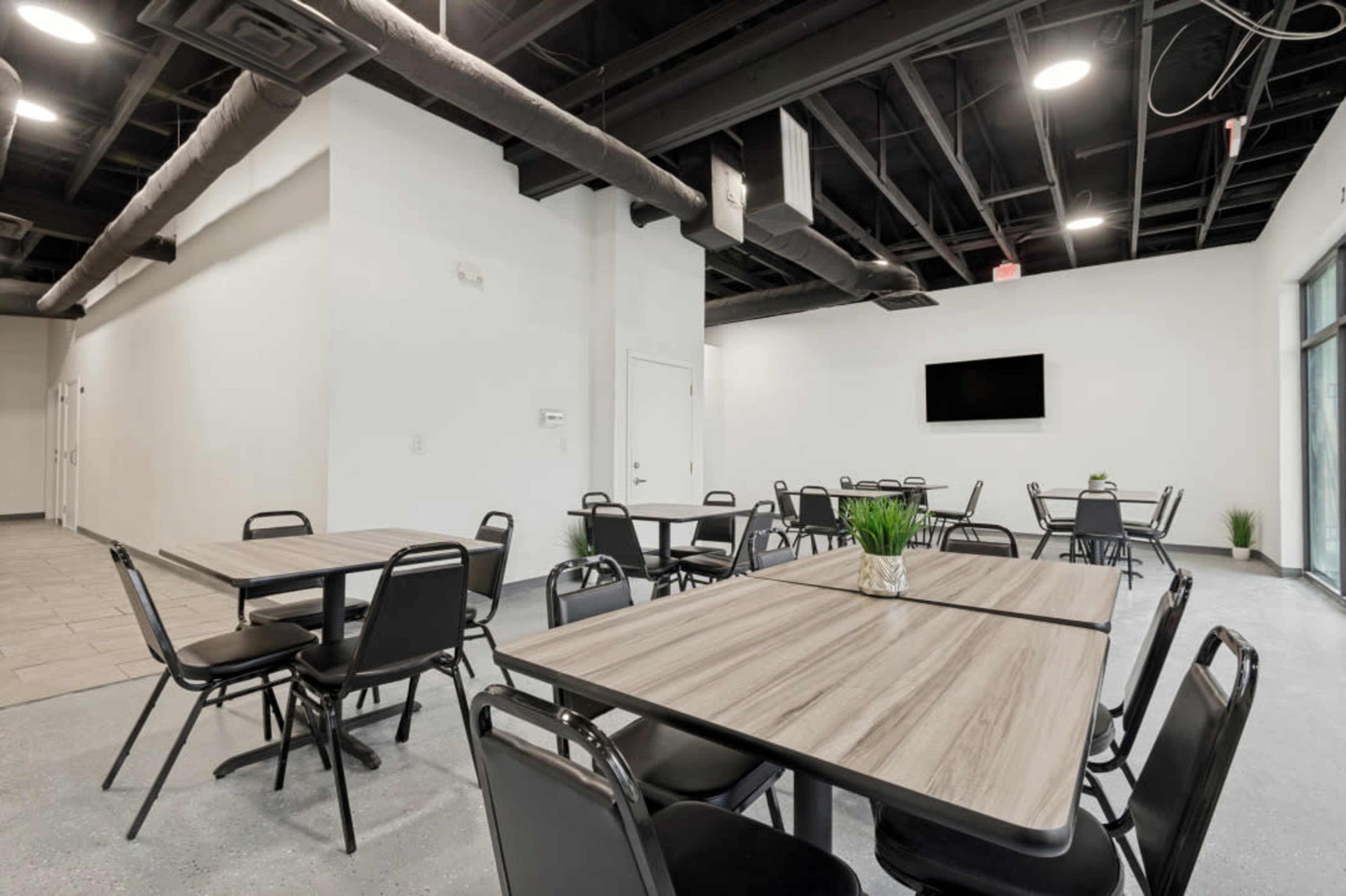 The image shows a modern, well-lit dining area featuring several rectangular tables and chairs arranged neatly against a plain white wall.