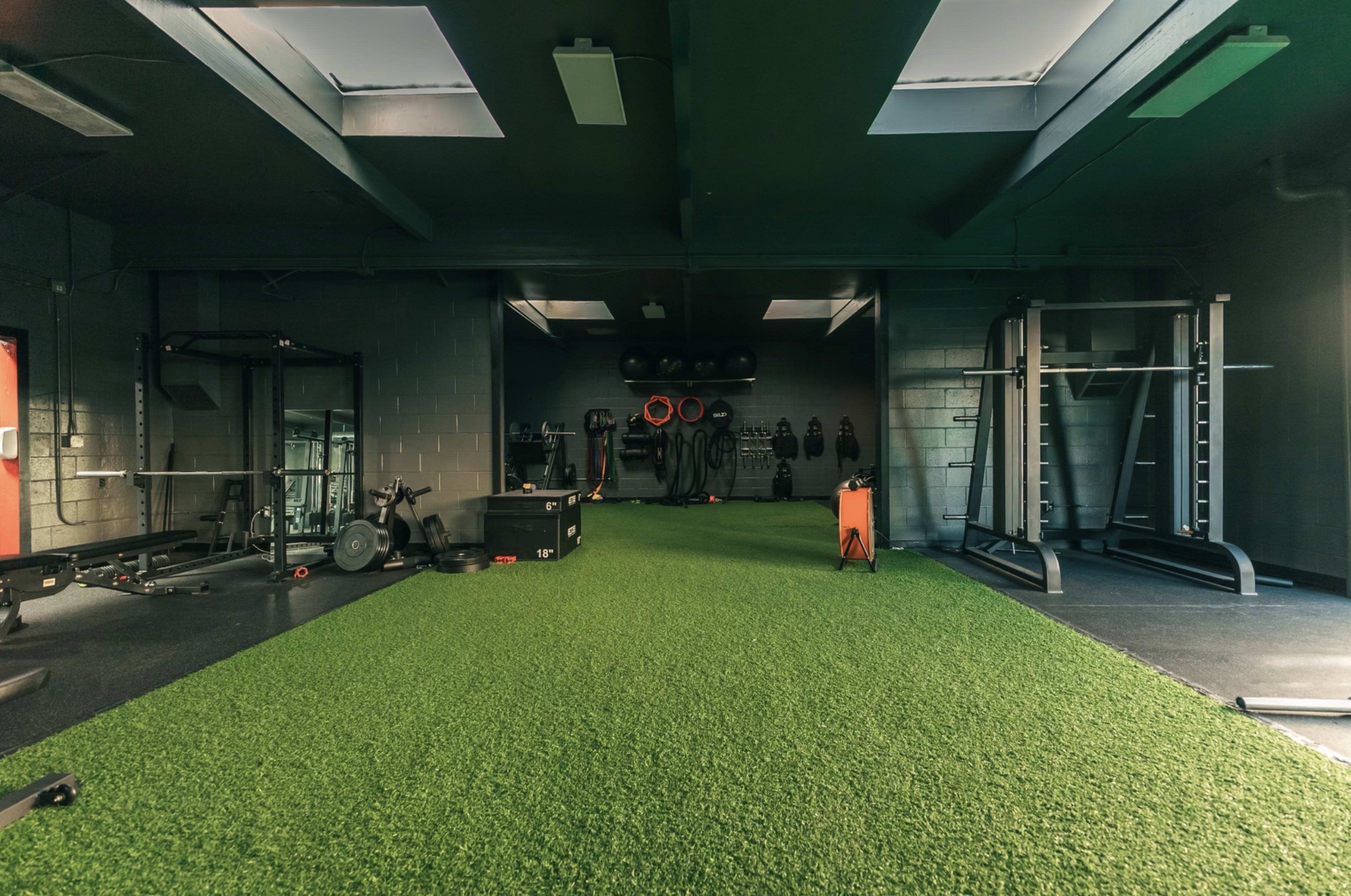 A fitness gym interior features artificial grass flooring, various exercise equipment, and weights arranged along the walls.