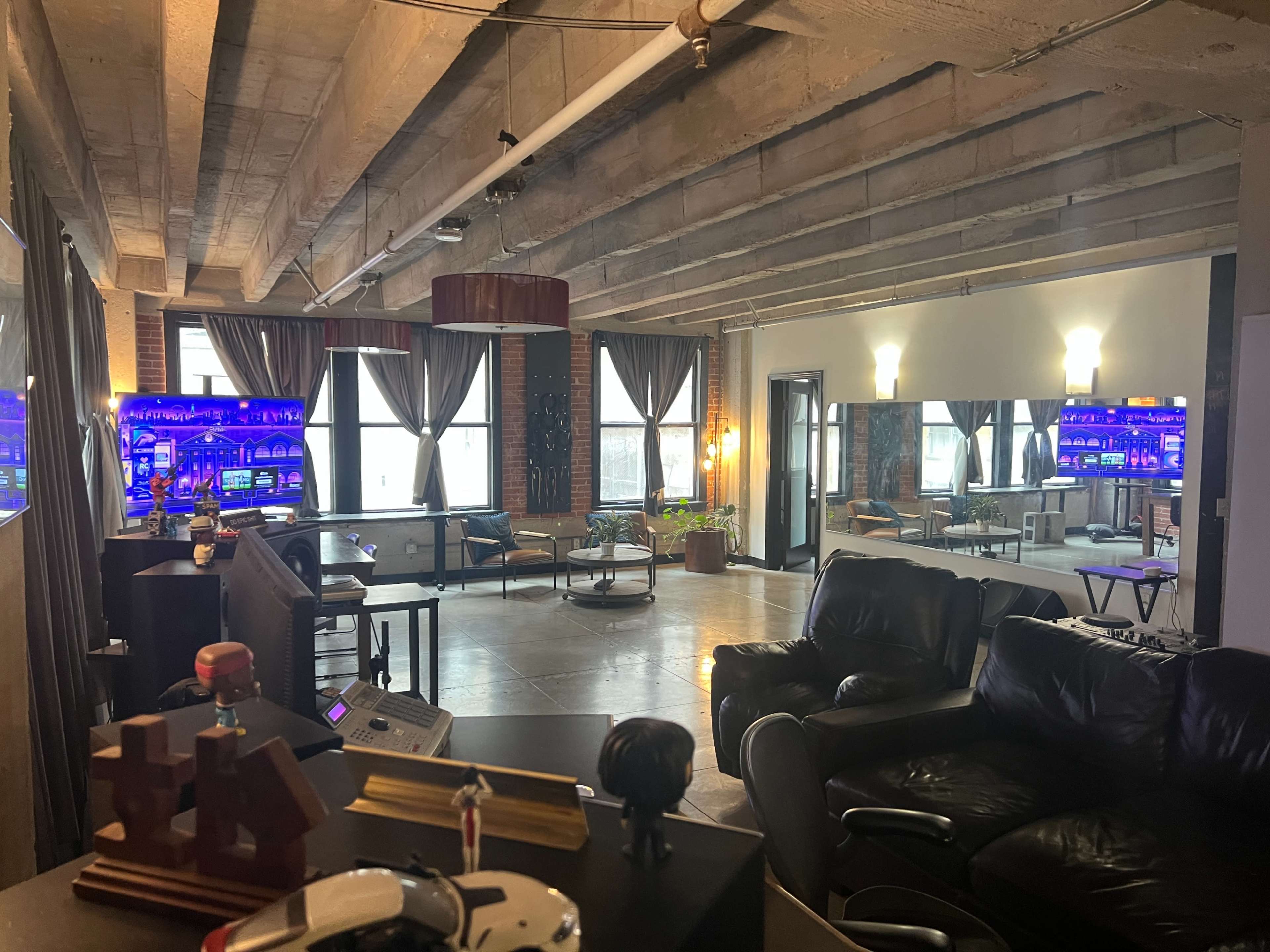 The image depicts a spacious, modern loft with exposed beams, large windows, and various seating areas, featuring a bar with ambient lighting in the background.