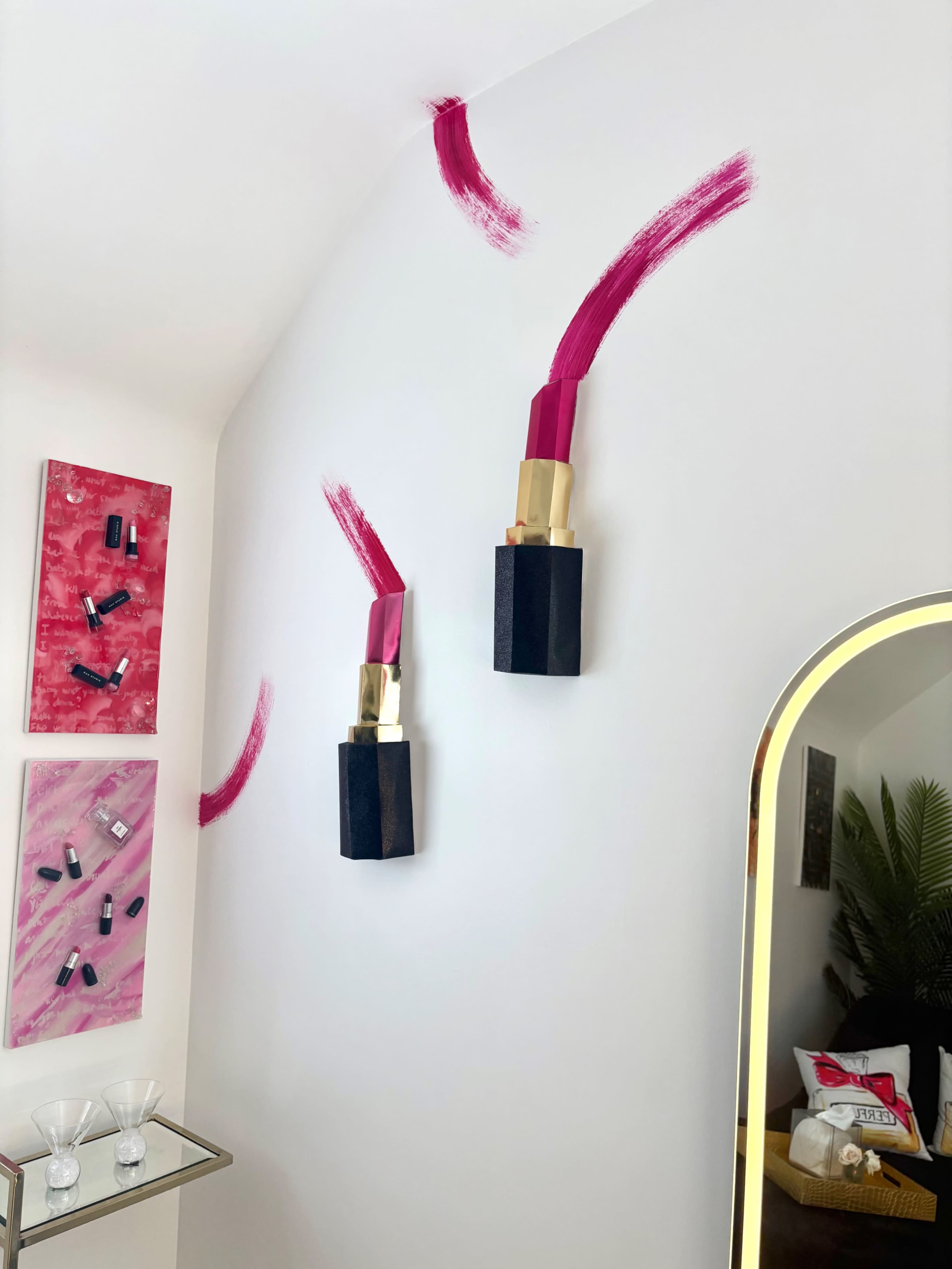 The image shows pink lipstick sculptures mounted on a white wall, accompanied by abstract pink and black artwork on the adjacent wall.