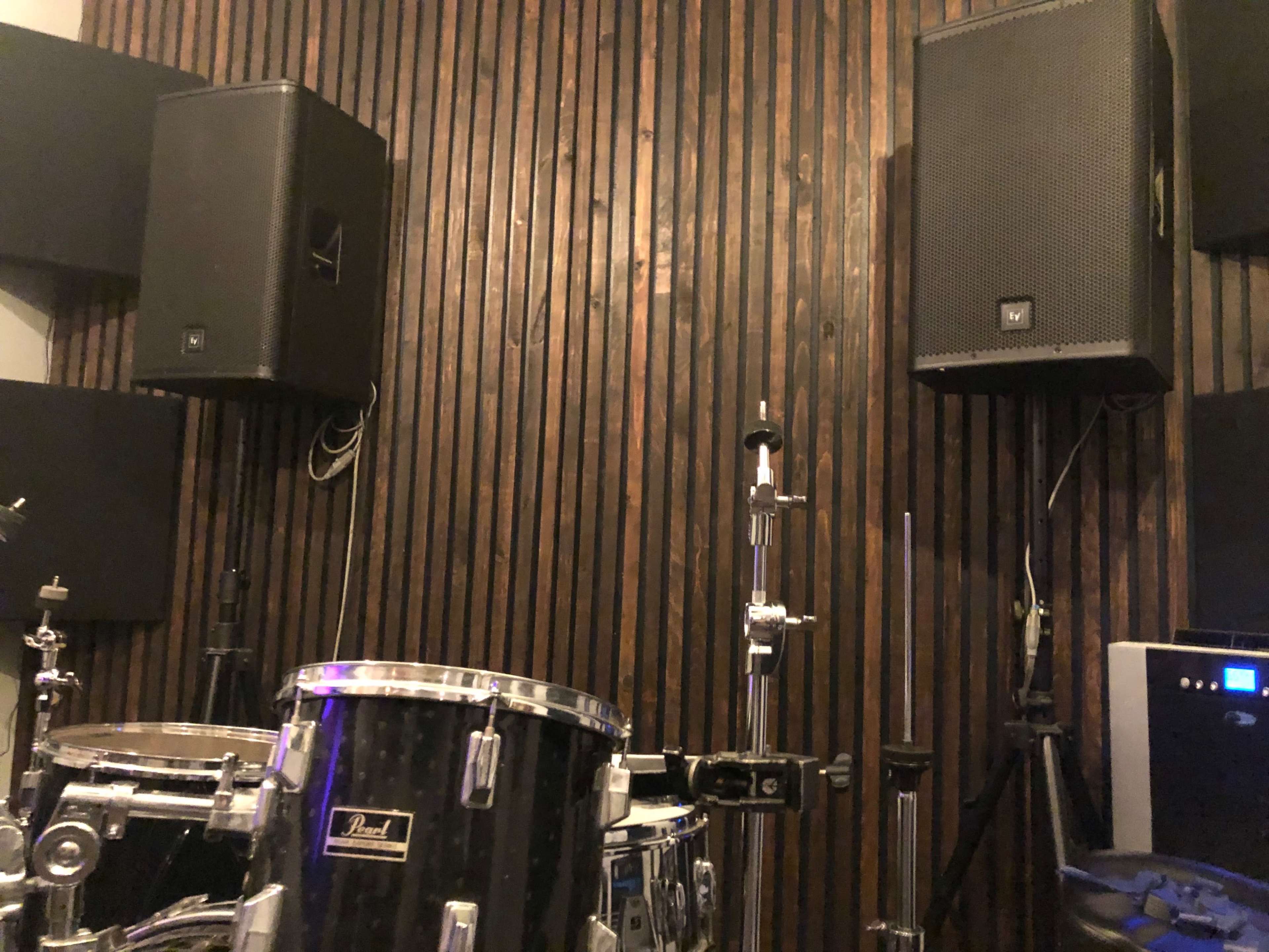 The image shows a wall with wooden paneling, flanked by speakers and a drum kit in a soundproof room.