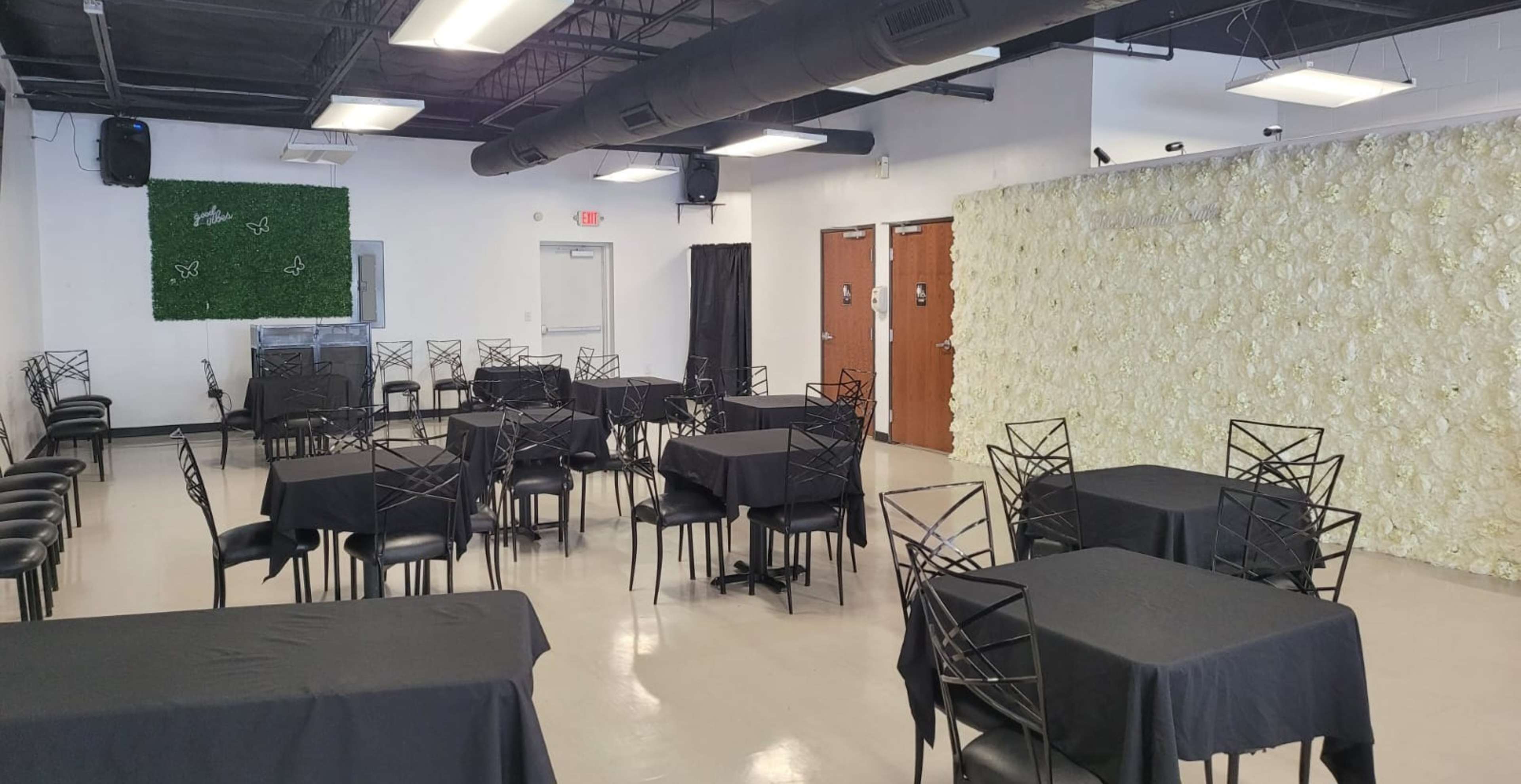 A room set up with black tablecloths on several tables and a wall adorned with artificial white flowers.