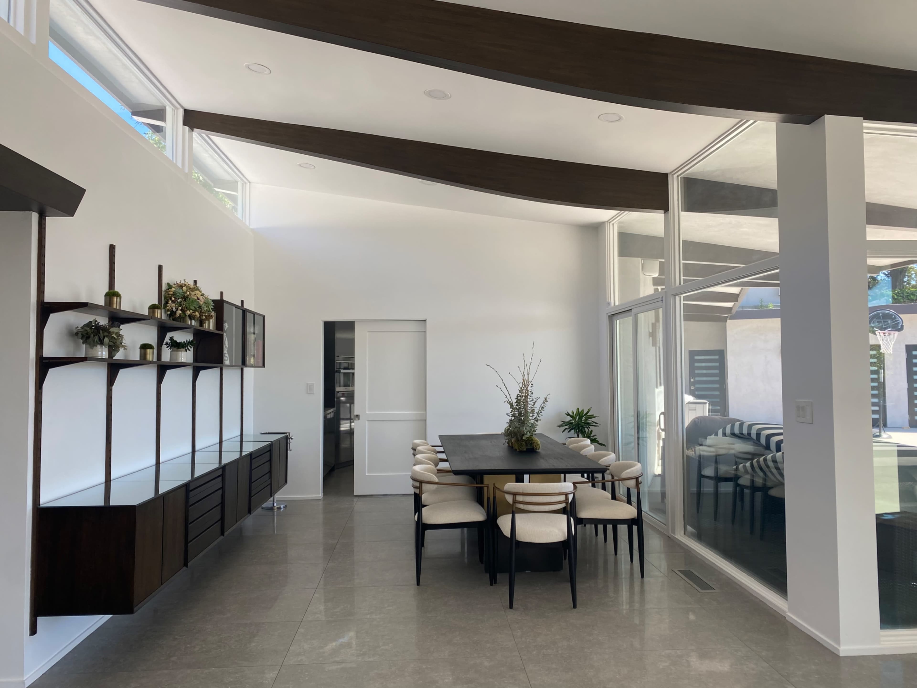 A modern dining area features a wooden table surrounded by upholstered chairs, with a sideboard and large windows allowing natural light.