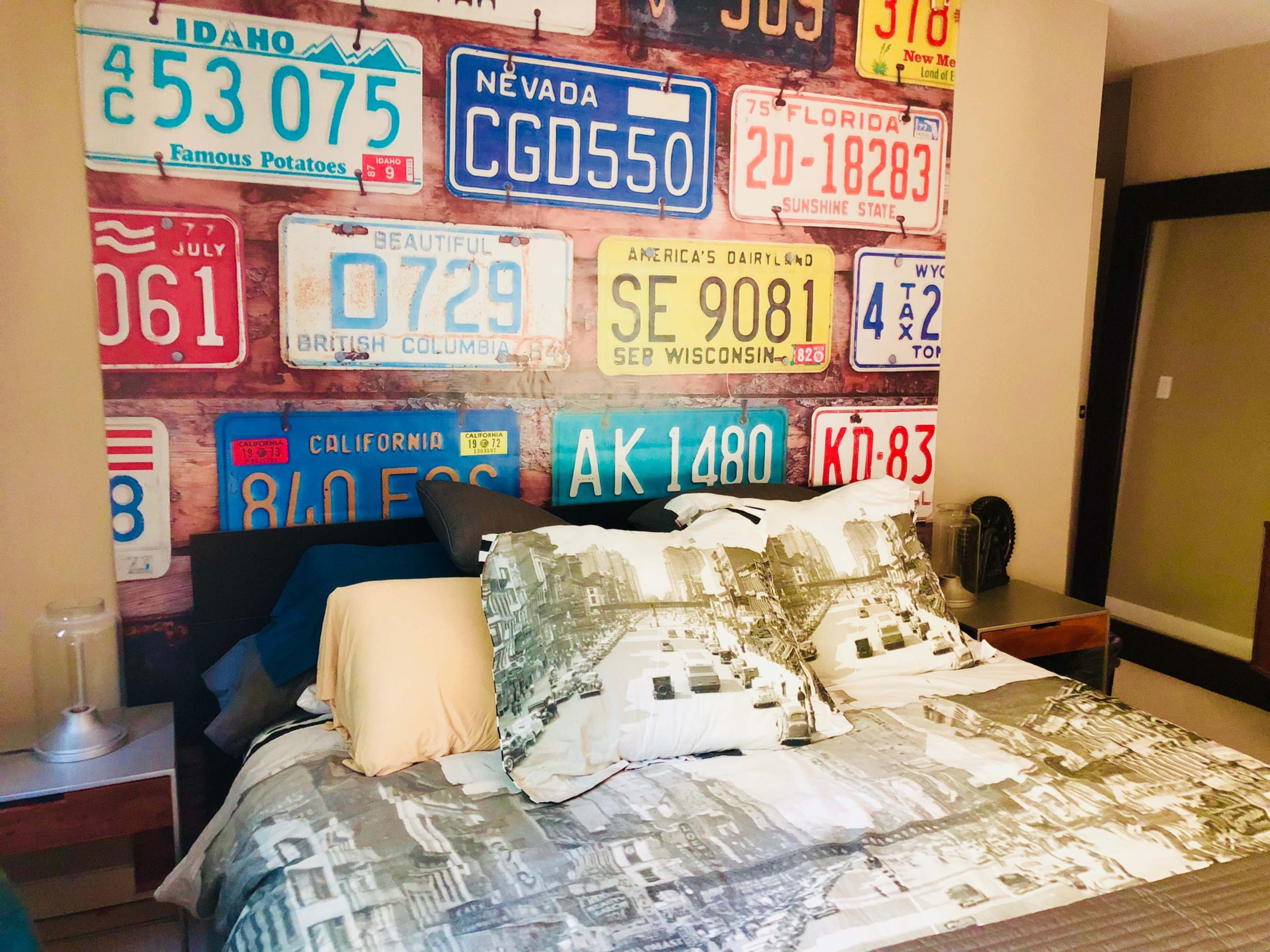 The image shows a bedroom with a wall mural made of various license plates and a bed with patterned pillows and a comforter featuring a cityscape.