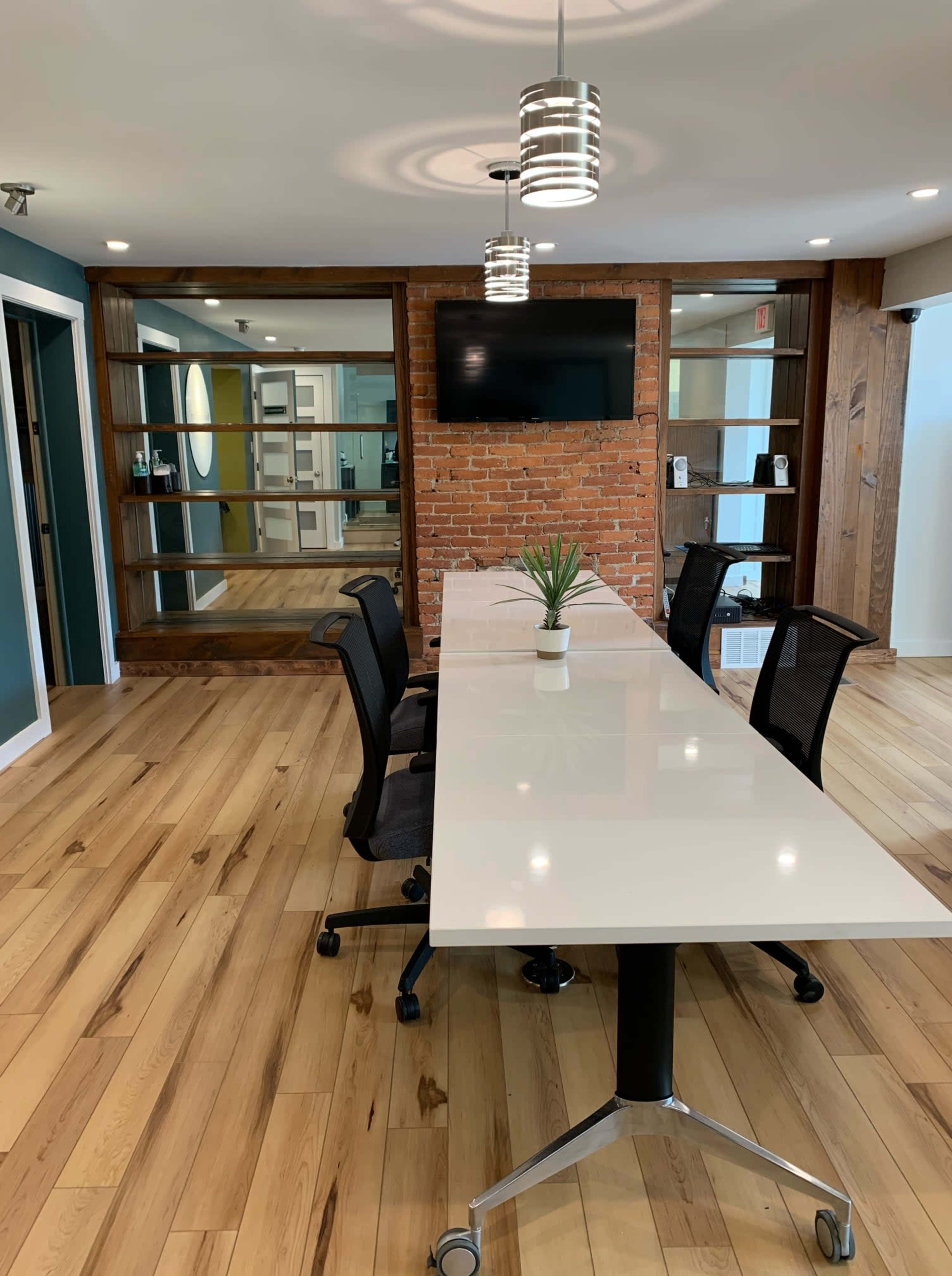 The image shows a modern conference room with a long white table, four black chairs, a television mounted on a brick wall, and glass shelving units.