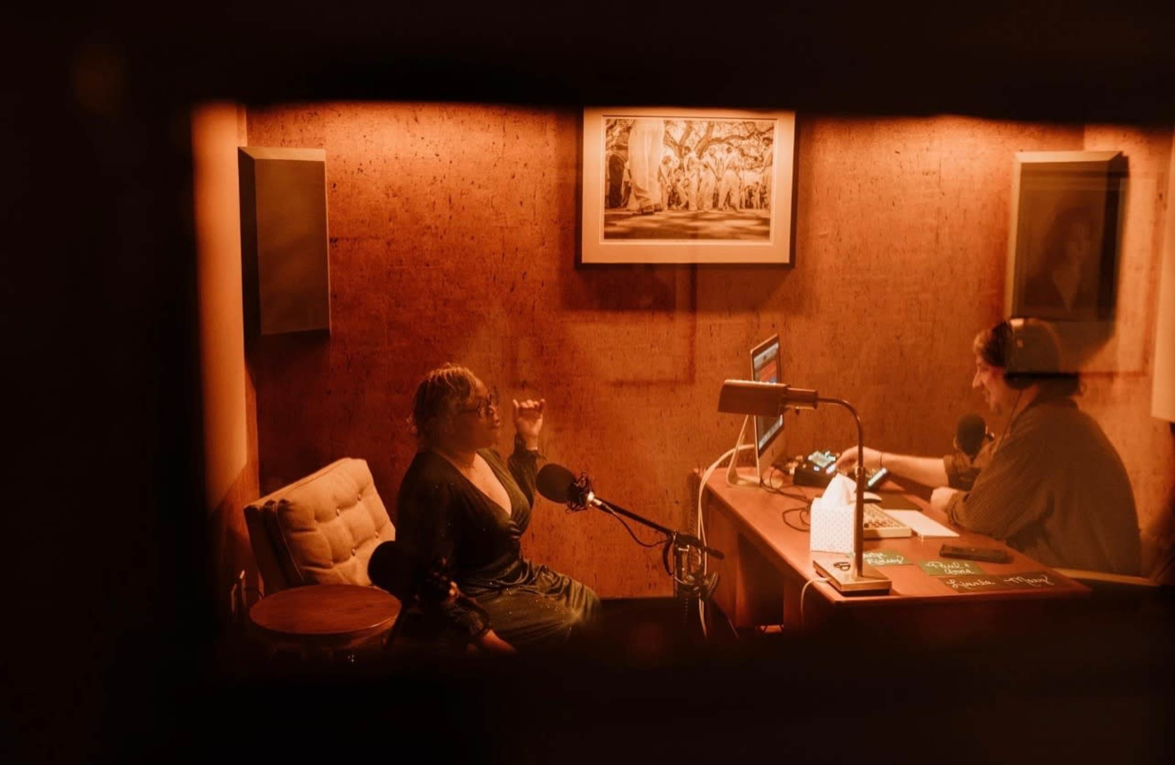 A woman sits in a recording studio speaking into a microphone while a man operates a computer and sound equipment across from her.