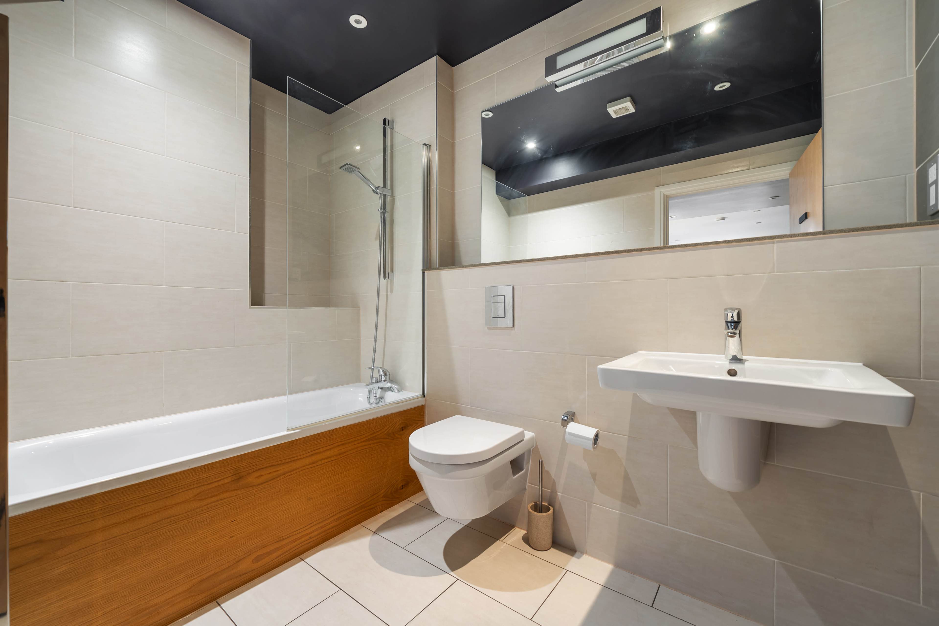 The image shows a modern bathroom featuring a bathtub with a shower, a wall-mounted sink, and a toilet, all set against light-colored tiled walls.