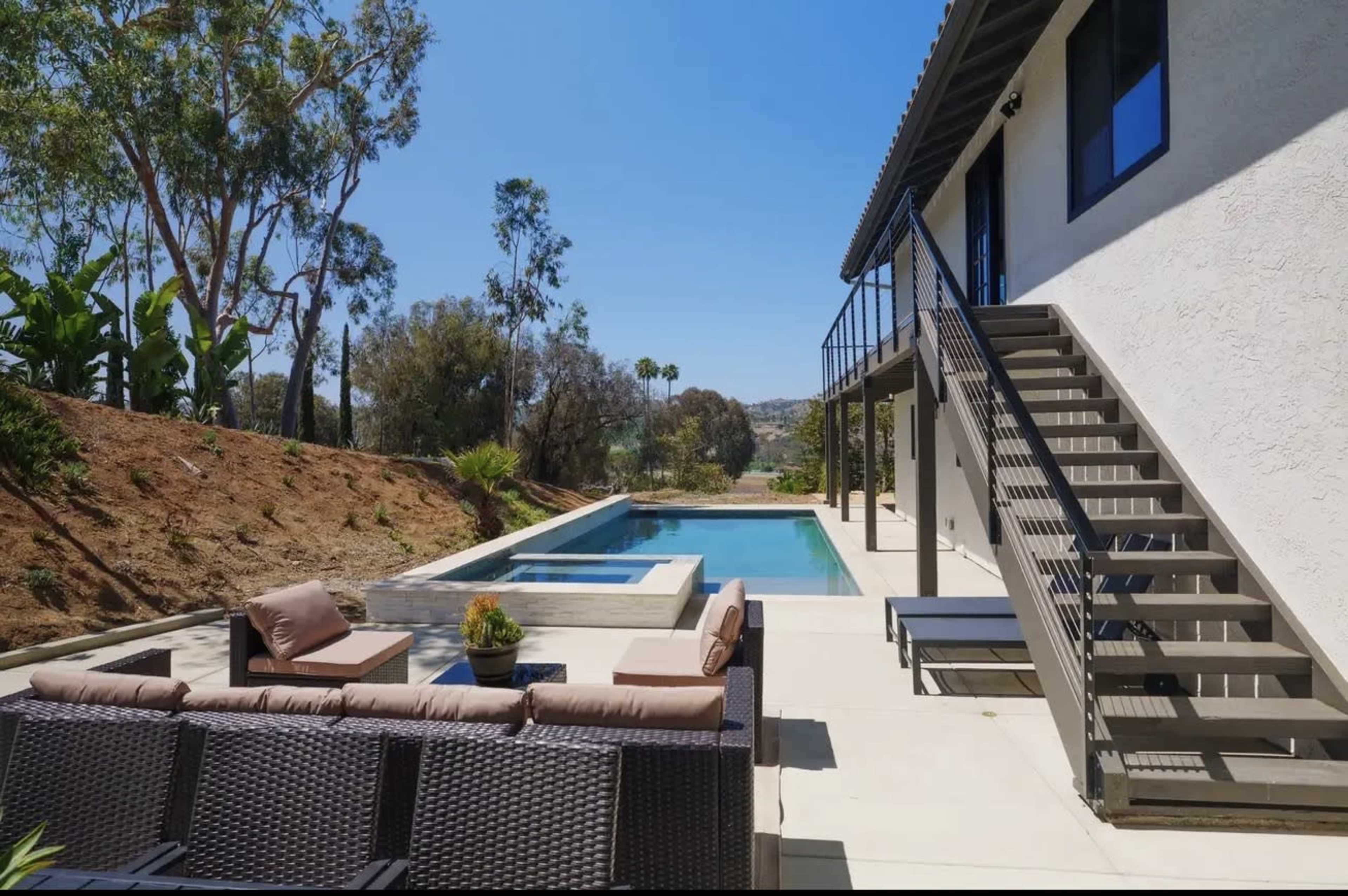 The image shows a modern outdoor living space featuring a lounge area and a swimming pool adjacent to a two-story house.