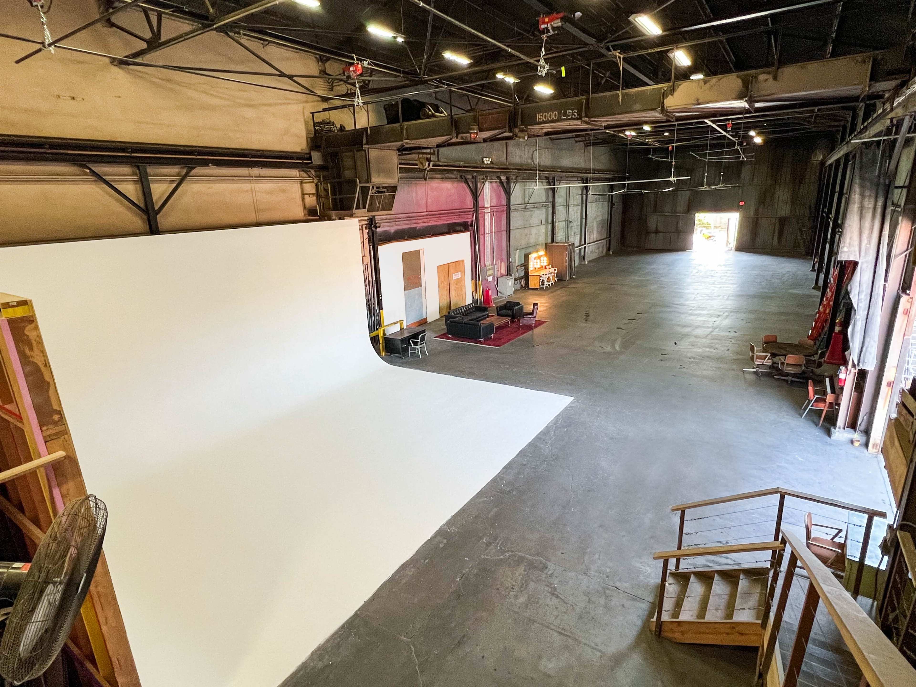 A spacious studio with a large white backdrop, industrial lighting, and various pieces of furniture in a well-lit area.
