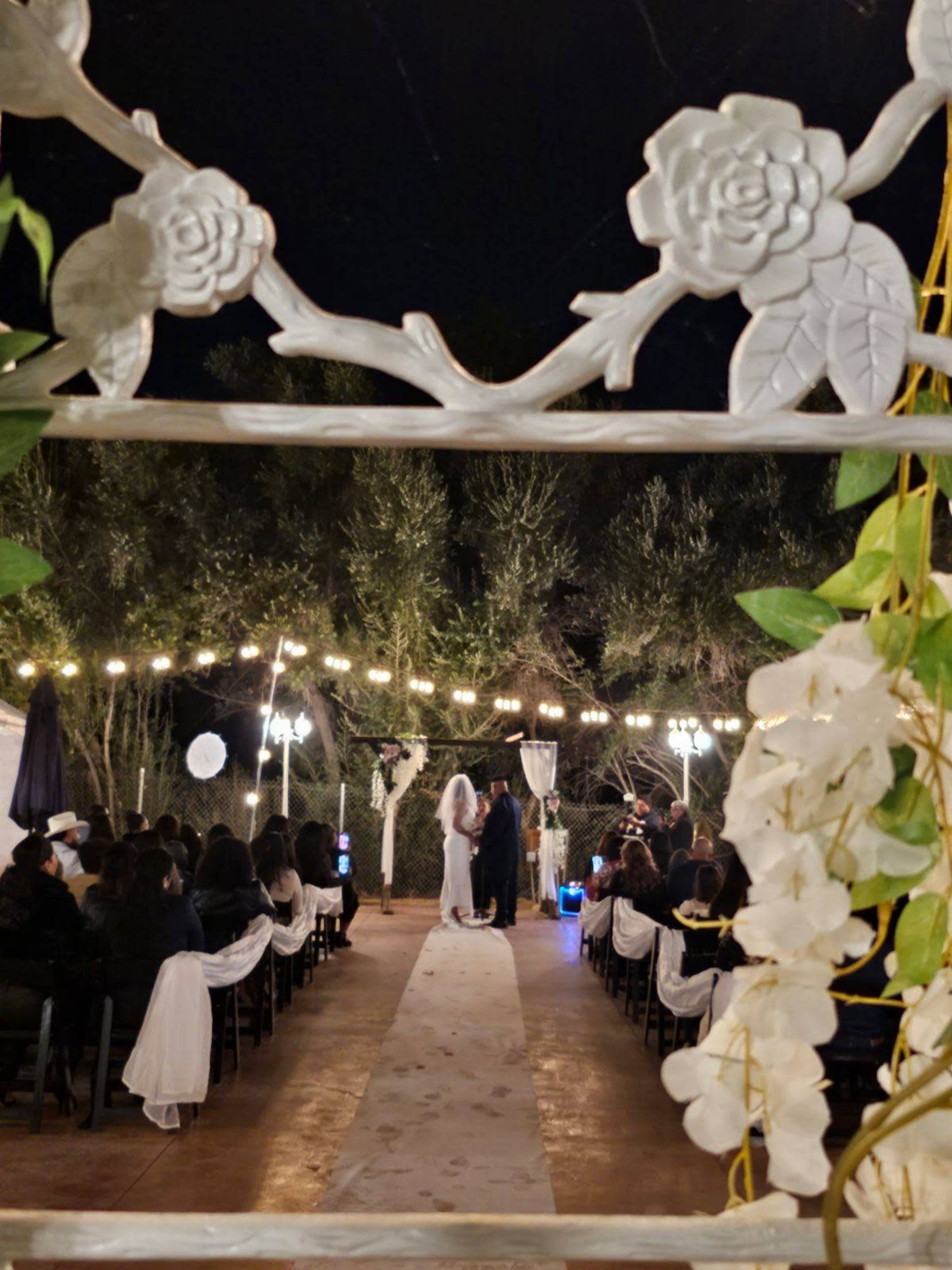A wedding ceremony is taking place outdoors under string lights, with guests seated on either side of a floral aisle.