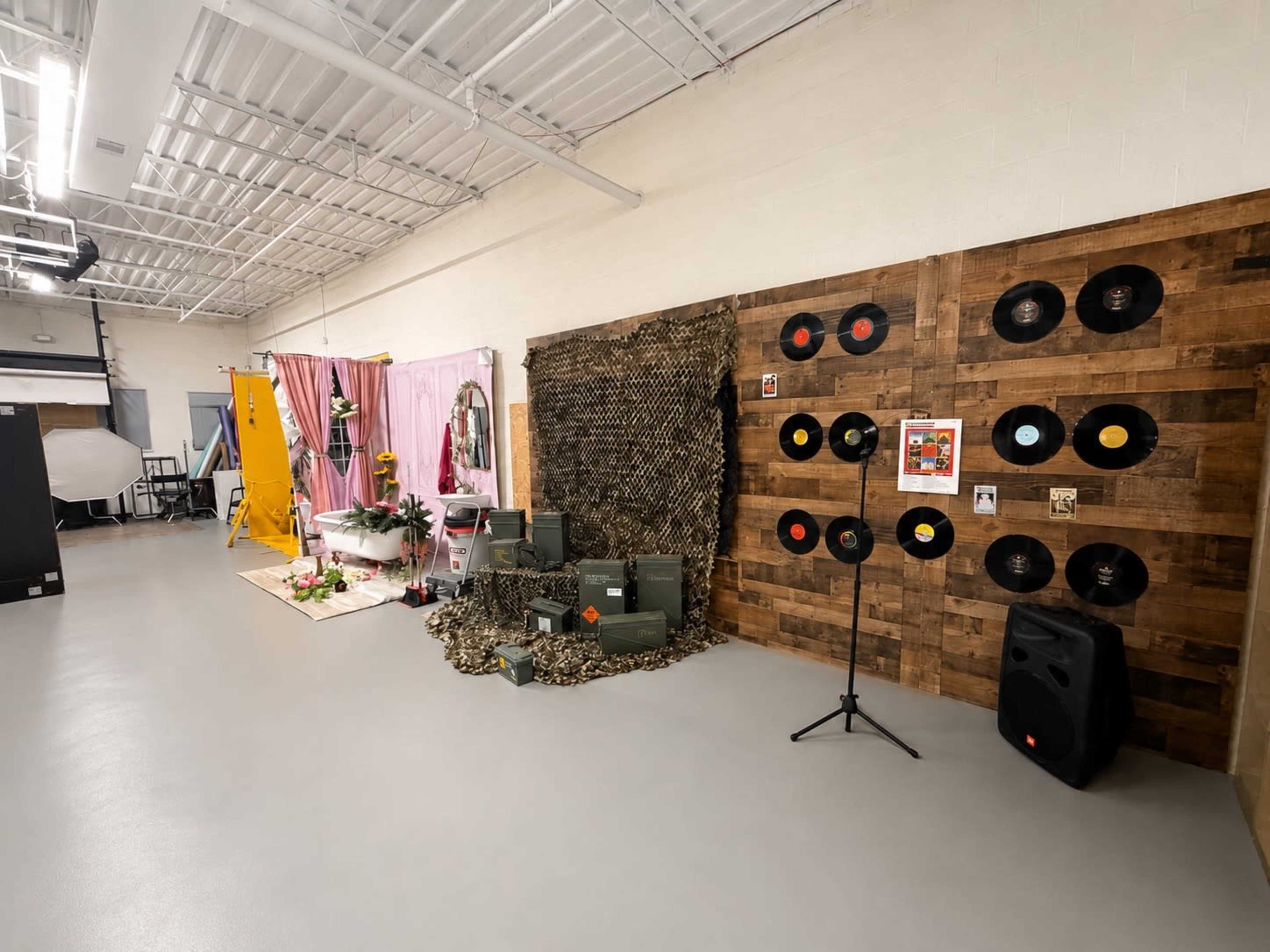 The image shows a spacious studio setup featuring a decorated wall with vinyl records, a camouflage display, and a floral arrangement in front of a backdrop.