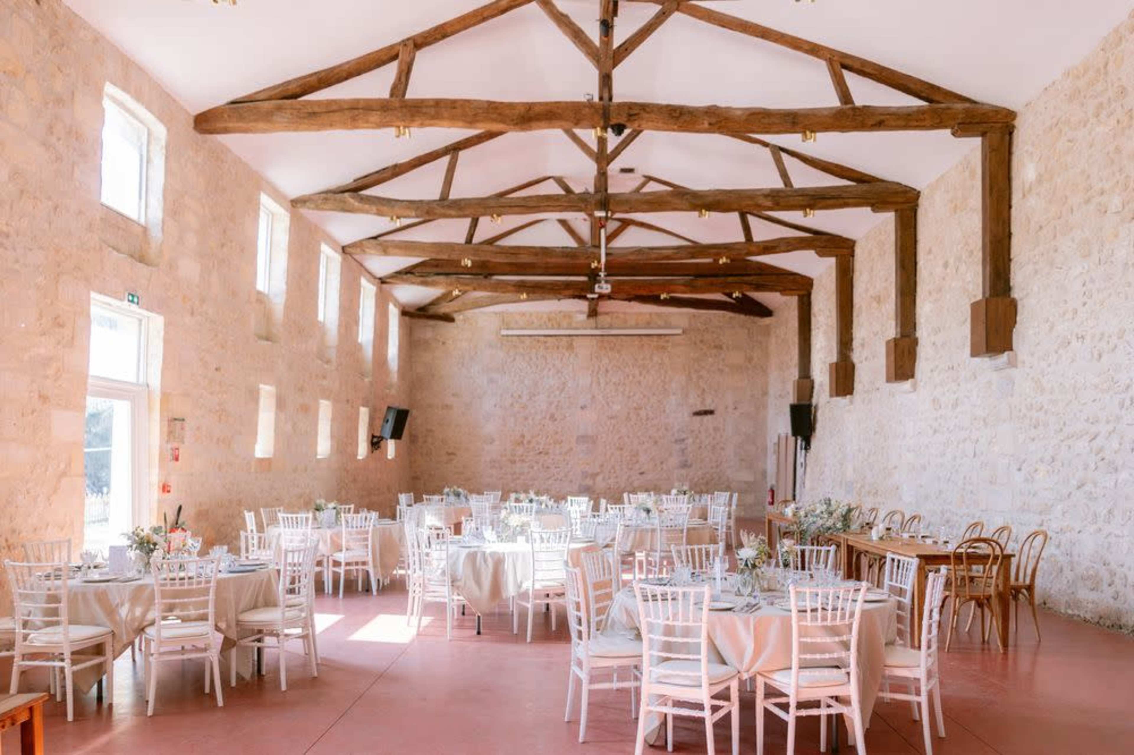 The image shows a spacious hall with stone walls and wooden beams, featuring several round tables set for dining and a long wooden table at the side.