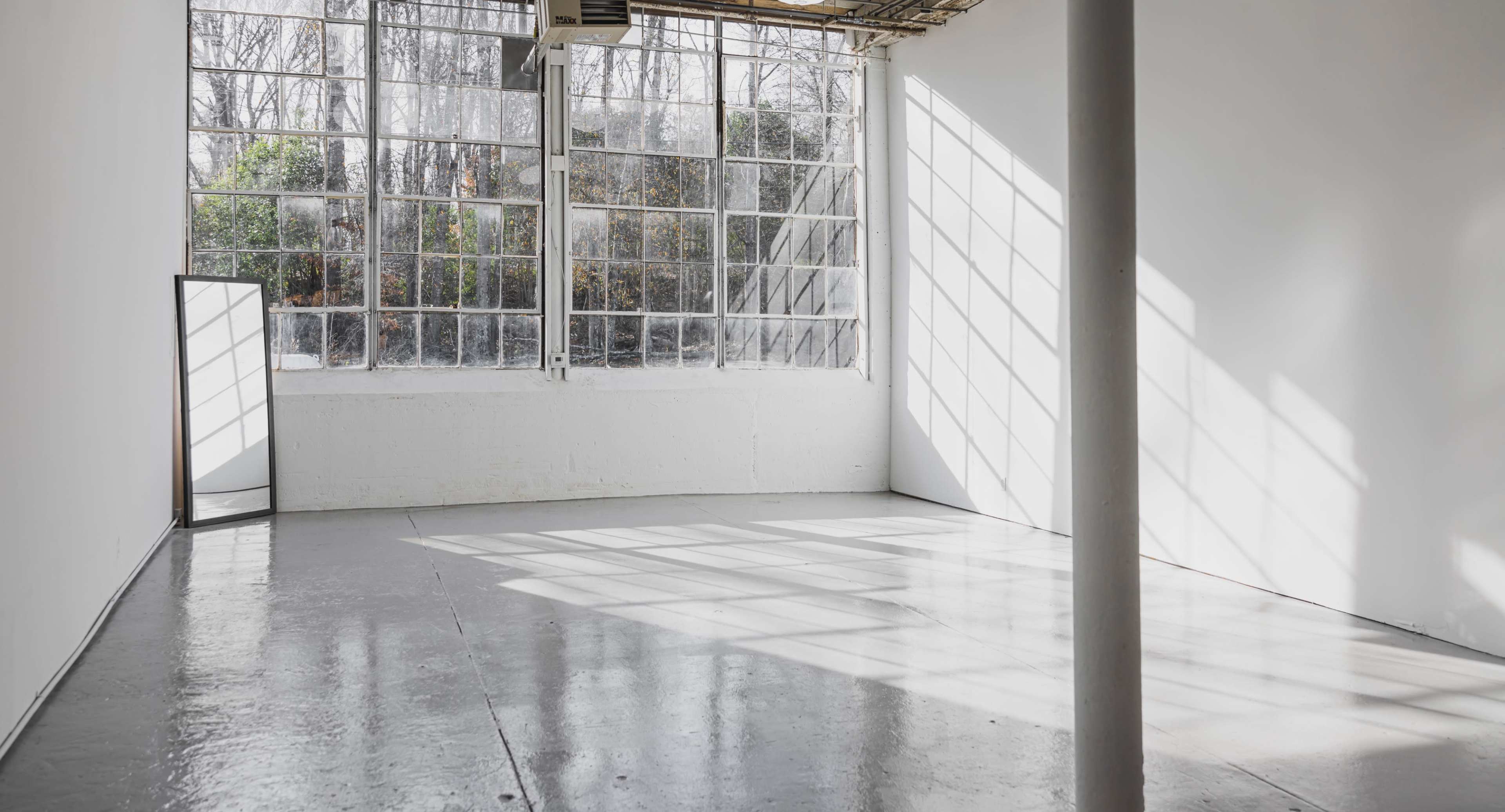 The image shows a spacious, empty room with large windows allowing sunlight to cast shadows on the polished concrete floor.