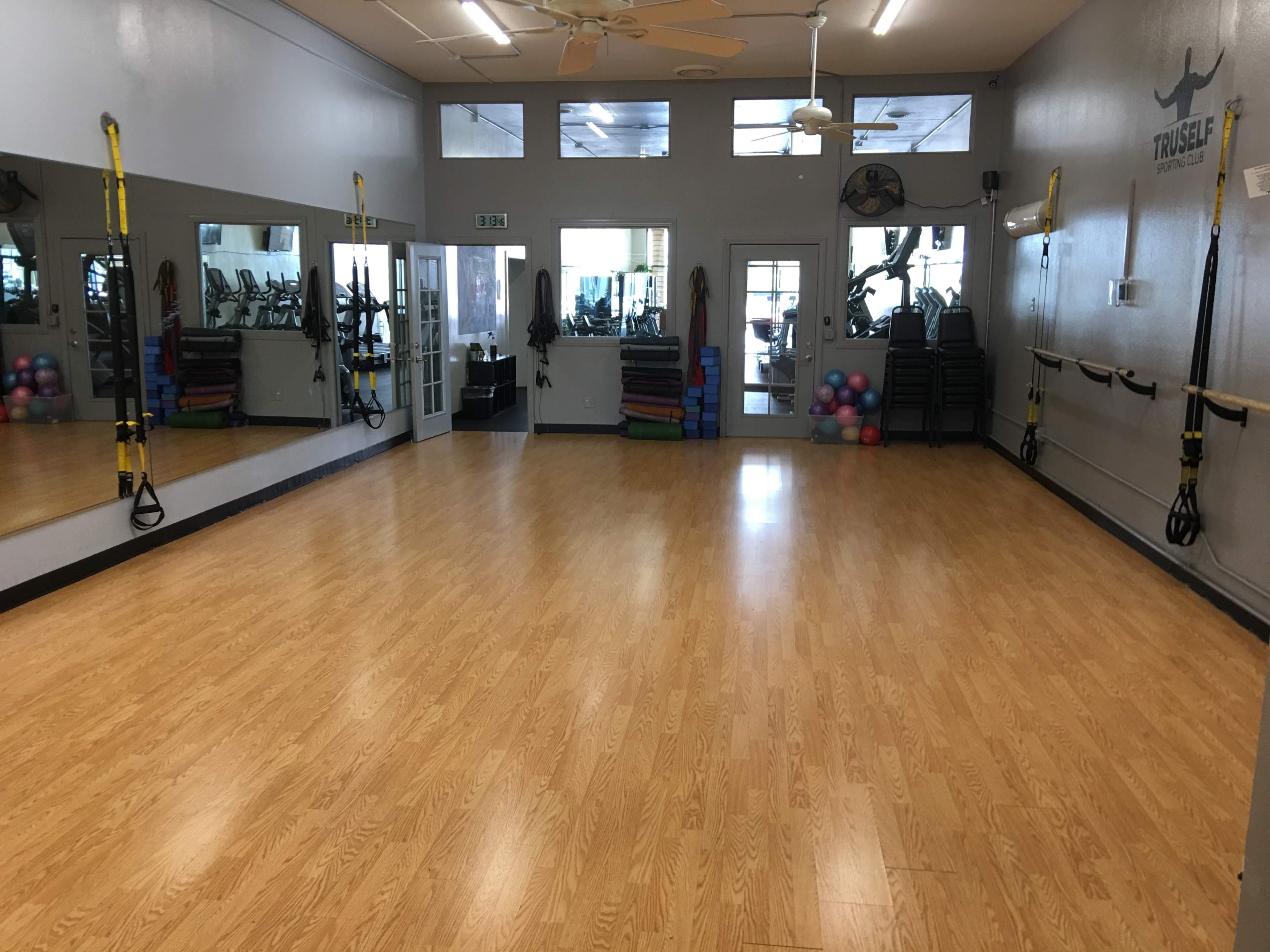 A spacious exercise studio with a wooden floor, large mirrors, and various fitness equipment along the walls.