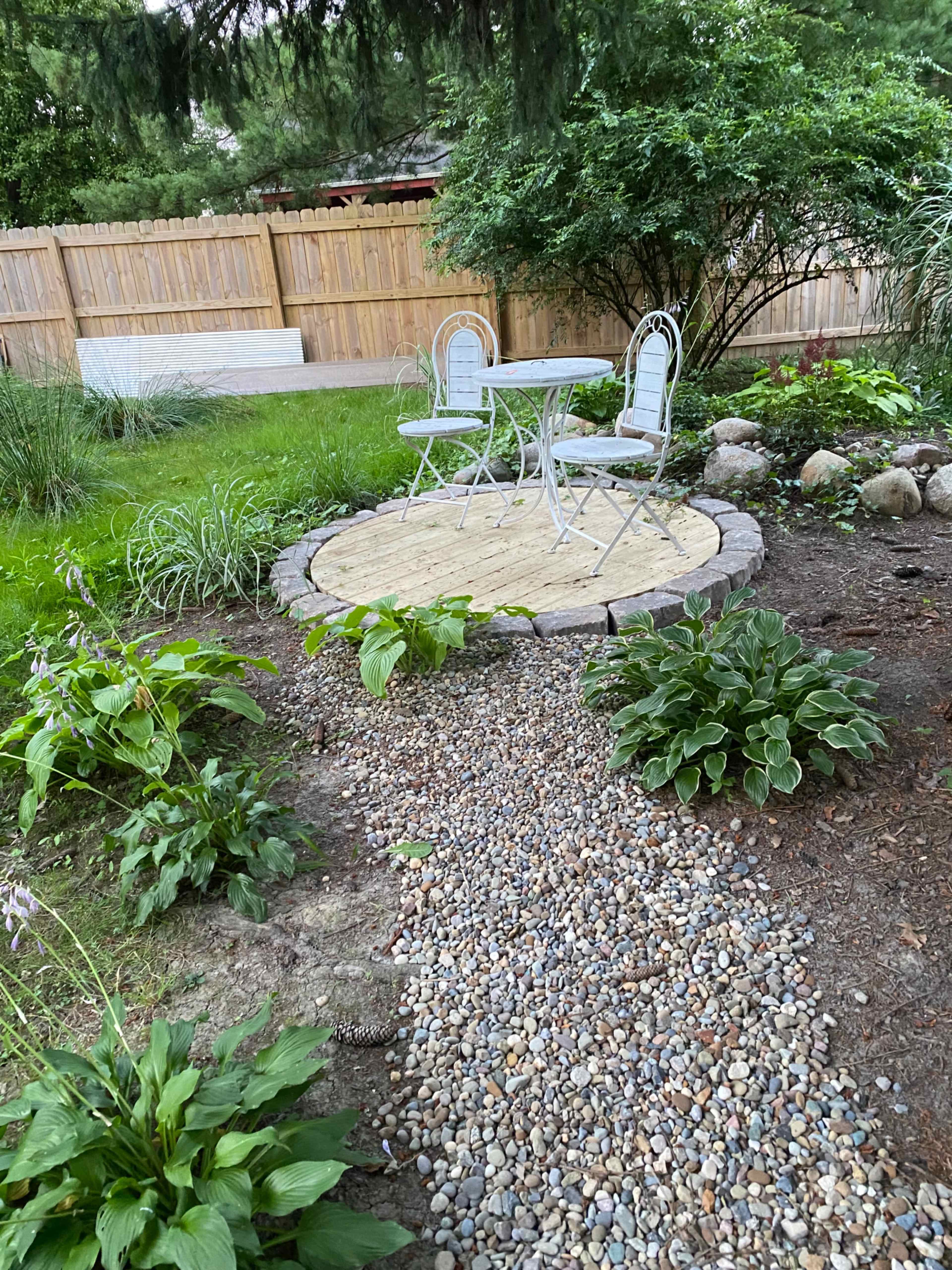 A circular patio made of wooden slats is surrounded by a stone border and two metal chairs, set in a garden with various plants and a gravel pathway.