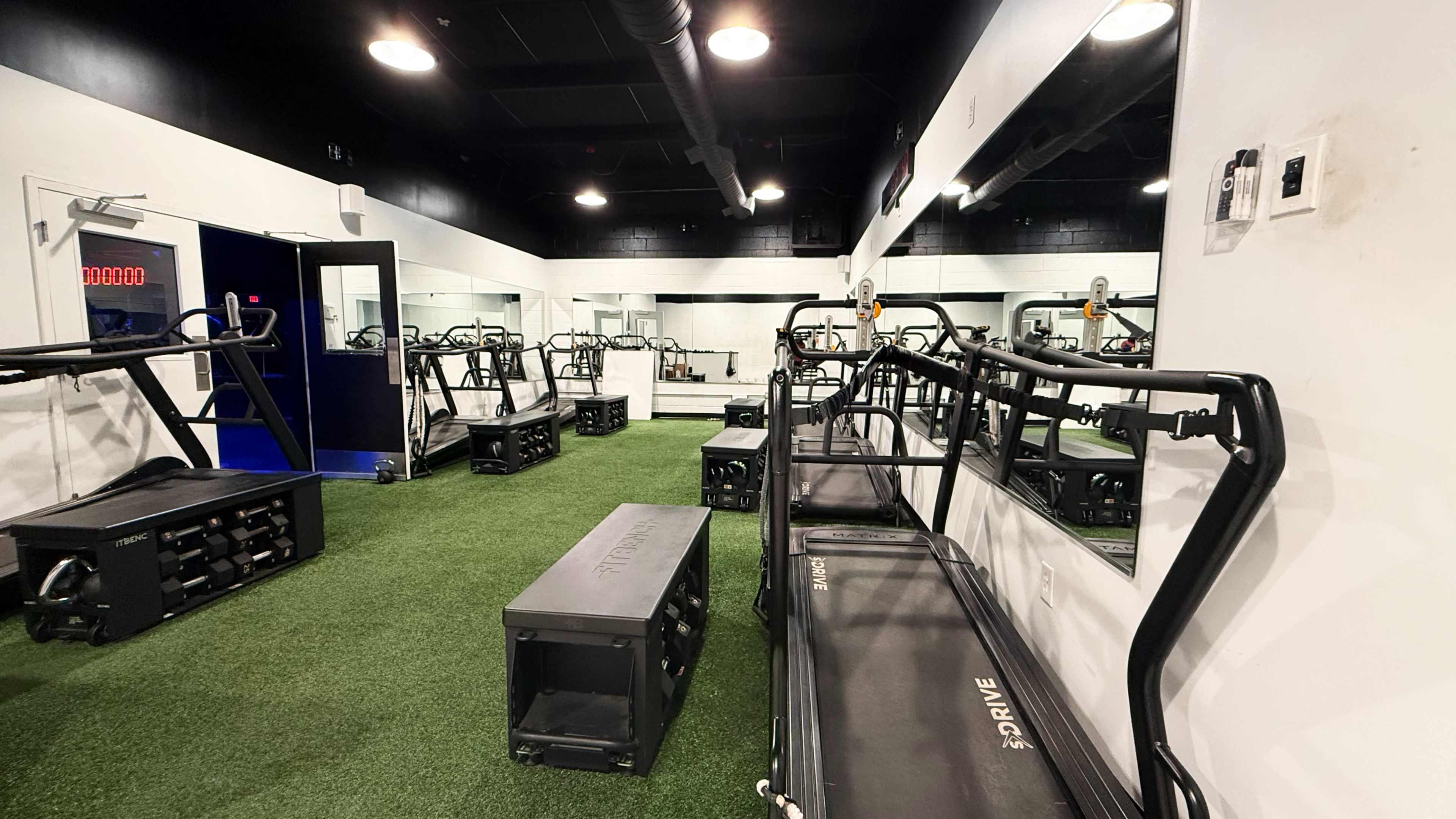 The image shows a fitness studio with several workout machines lined up on a green turf floor, along with mirrors on the walls.