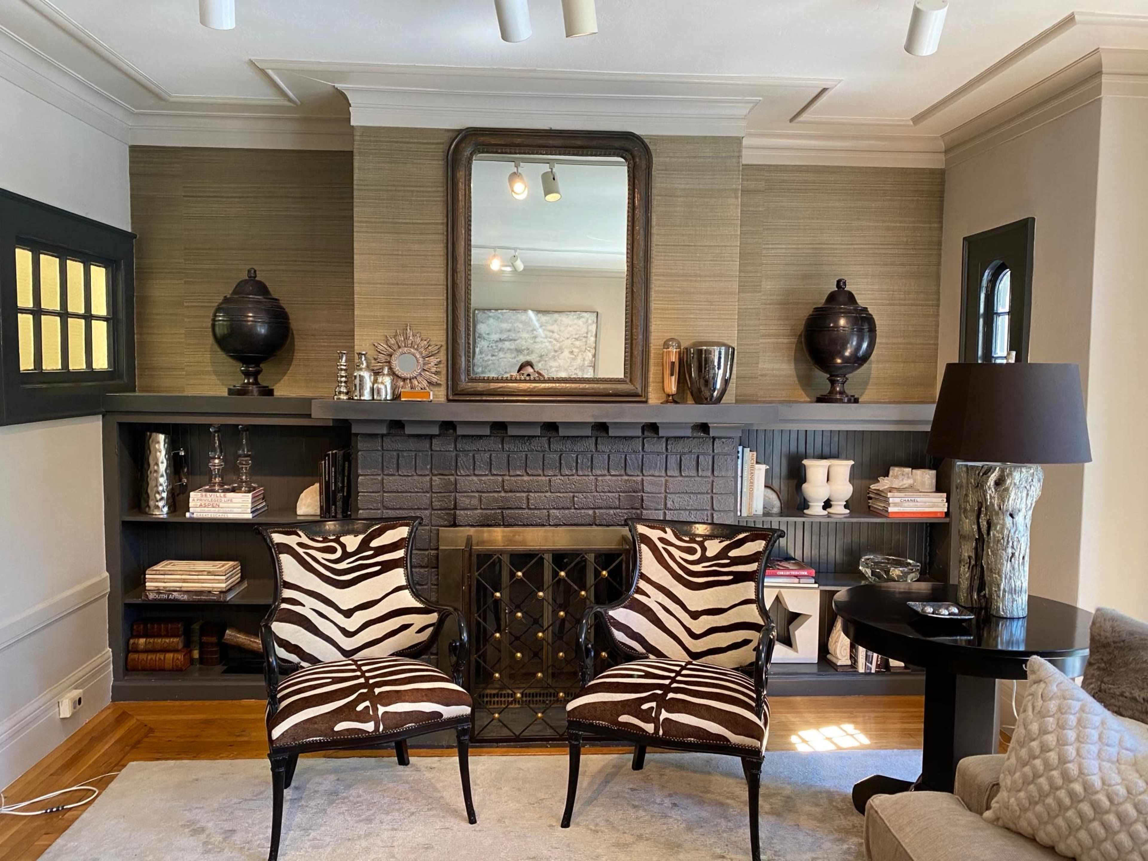 The image shows a living room with two zebra-striped chairs facing a black brick fireplace, flanked by decorative vases and a large mirror above the mantel.