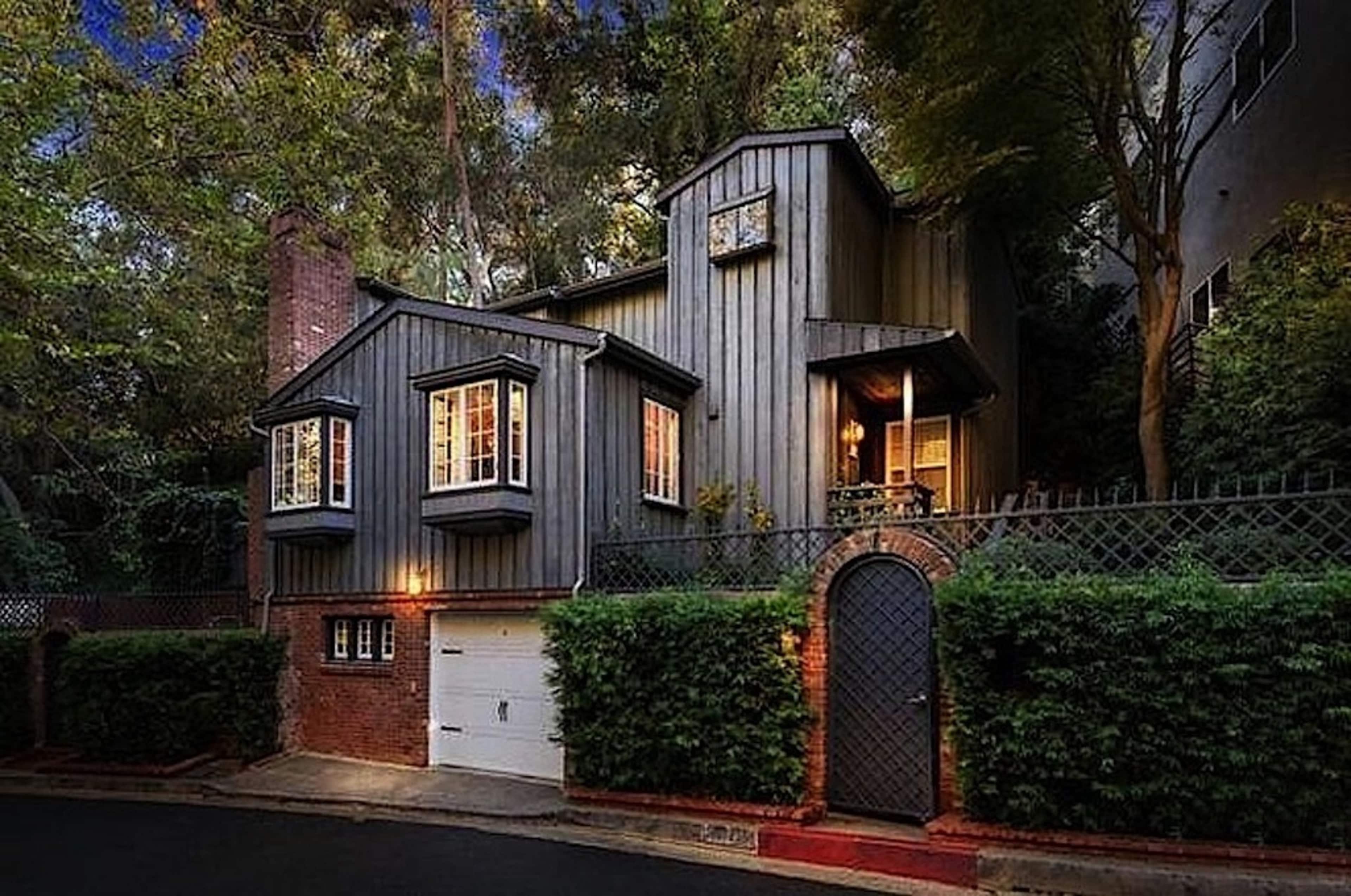 A two-story wooden house with large windows and a brick garage is surrounded by trees and greenery.