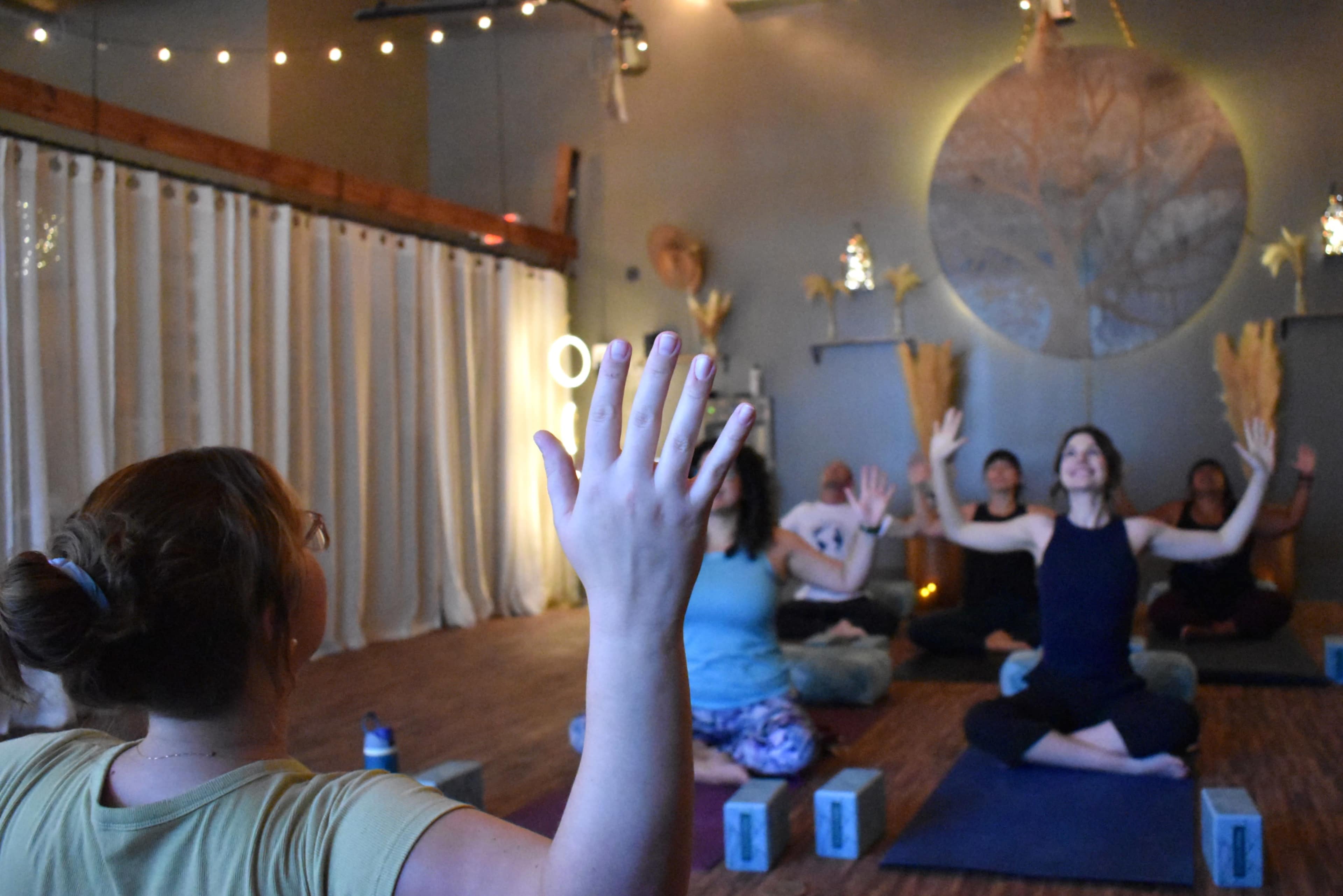 A group of people are seated in a yoga studio, practicing poses and raising their arms while facing a teacher at the front.