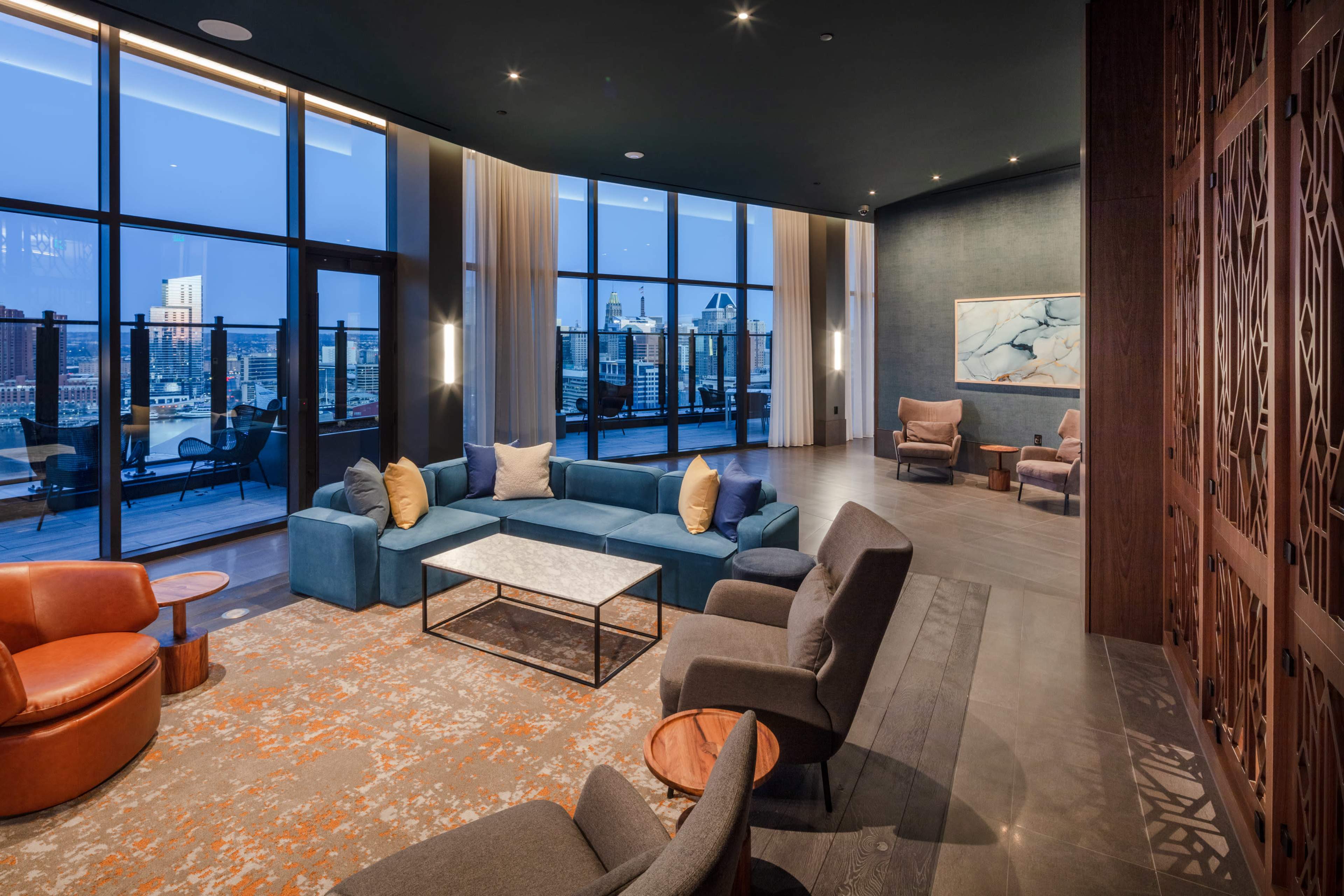 The image shows a modern lounge area with a blue sofa, armchairs, and a coffee table, featuring large windows that overlook a city skyline at dusk.