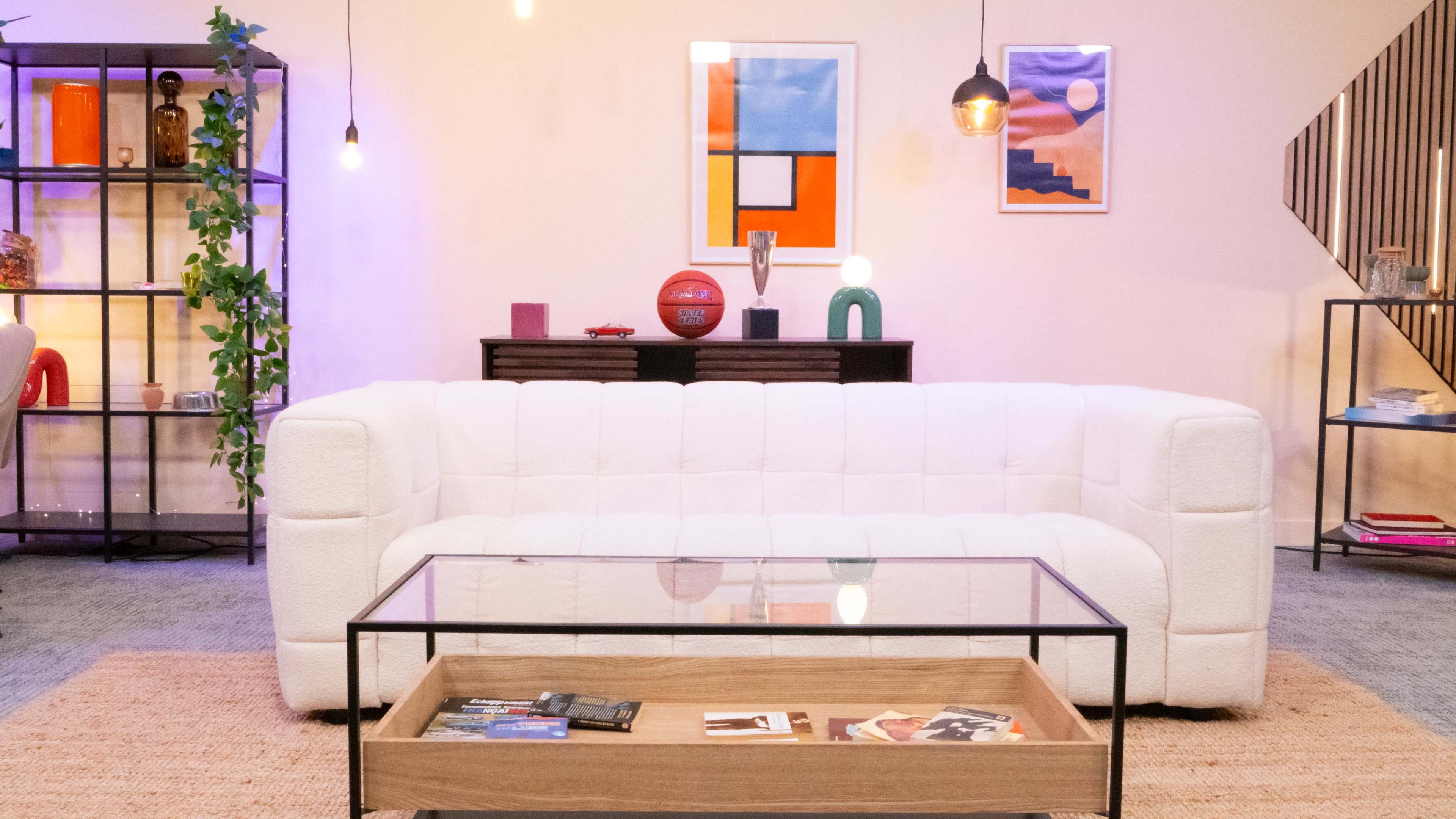The image shows a cozy living room setting featuring a white tufted sofa, a glass coffee table with magazines, and colorful artwork on the wall.