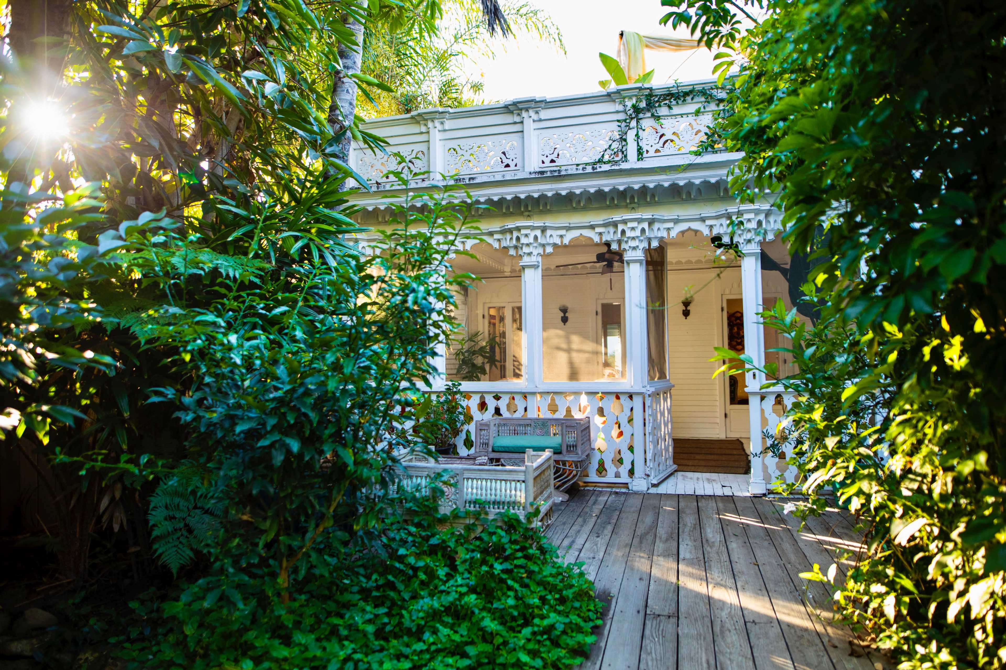 A white, Victorian-style house is nestled among lush greenery, with a wooden deck leading to its entrance.