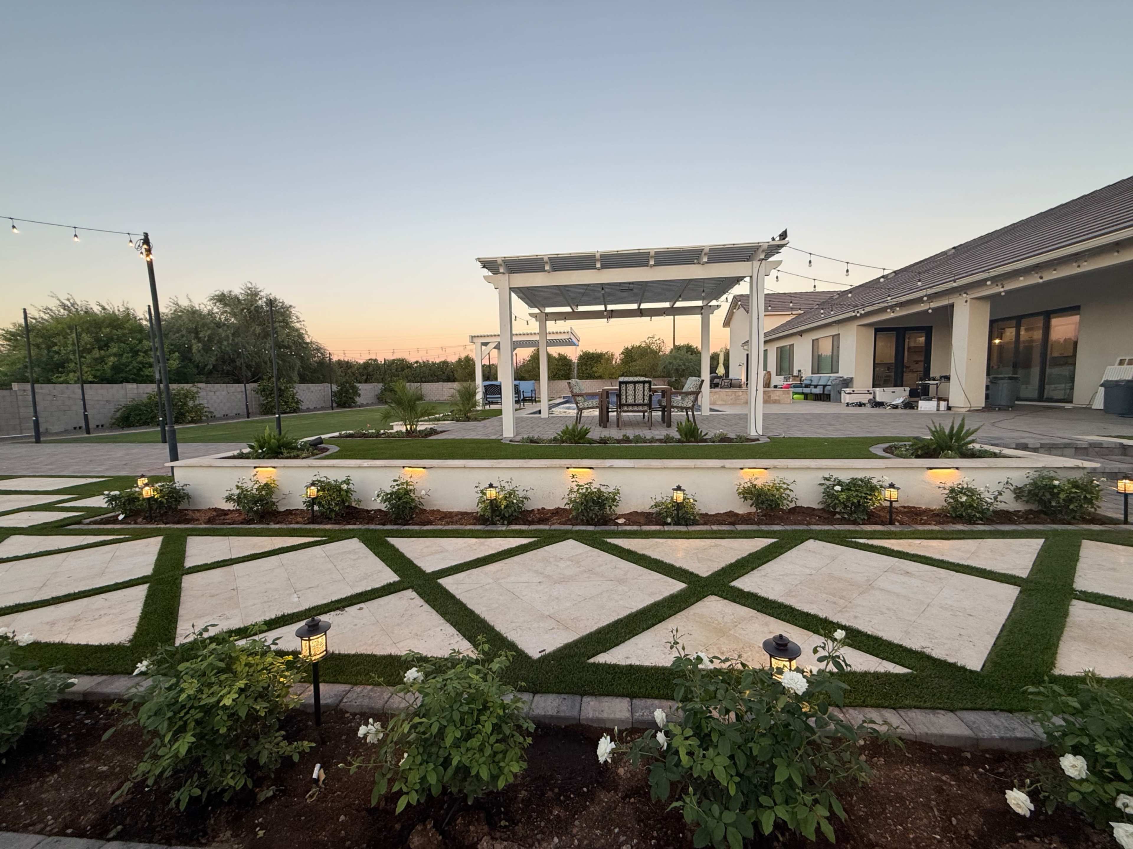 The image shows a landscaped backyard featuring a gazebo, seating area, symmetrical garden beds, and pathway lights, all set against a clear sky at dusk.