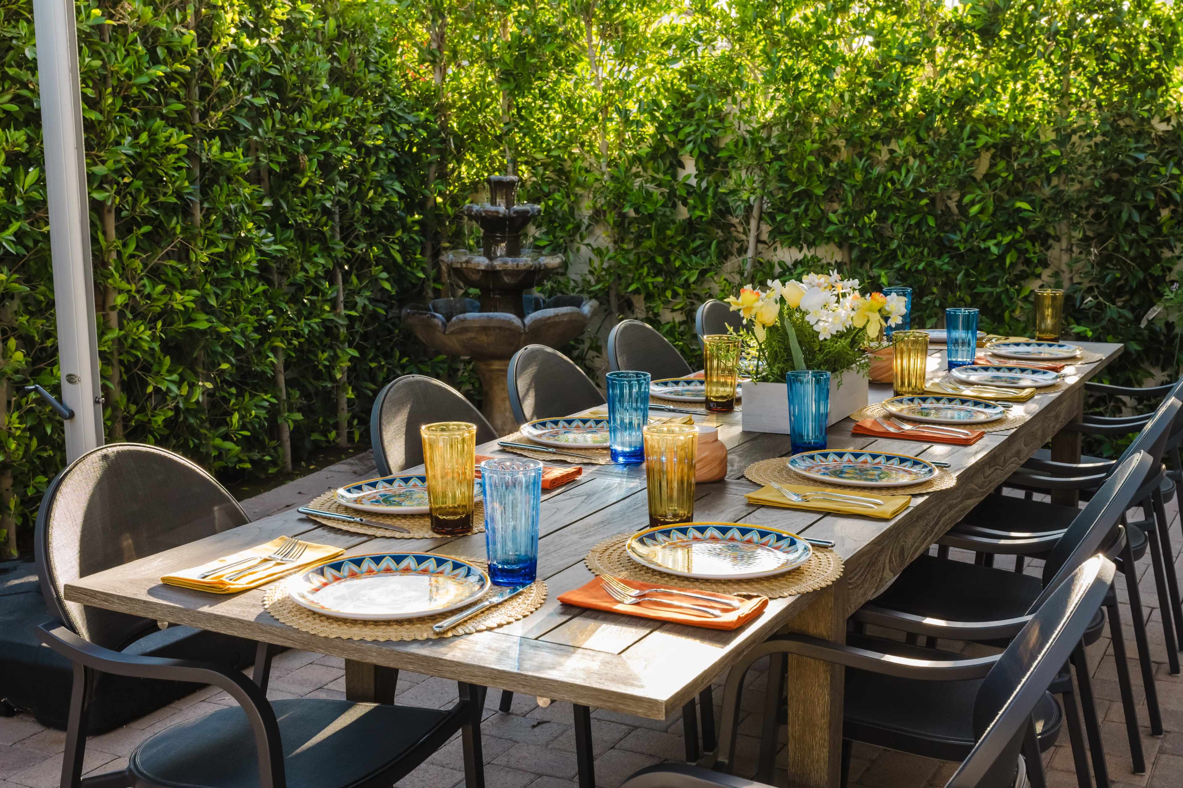 A long wooden dining table is set for a meal outdoors, adorned with colorful plates, glasses, and a floral centerpiece, with a green hedge and a fountain in the background.