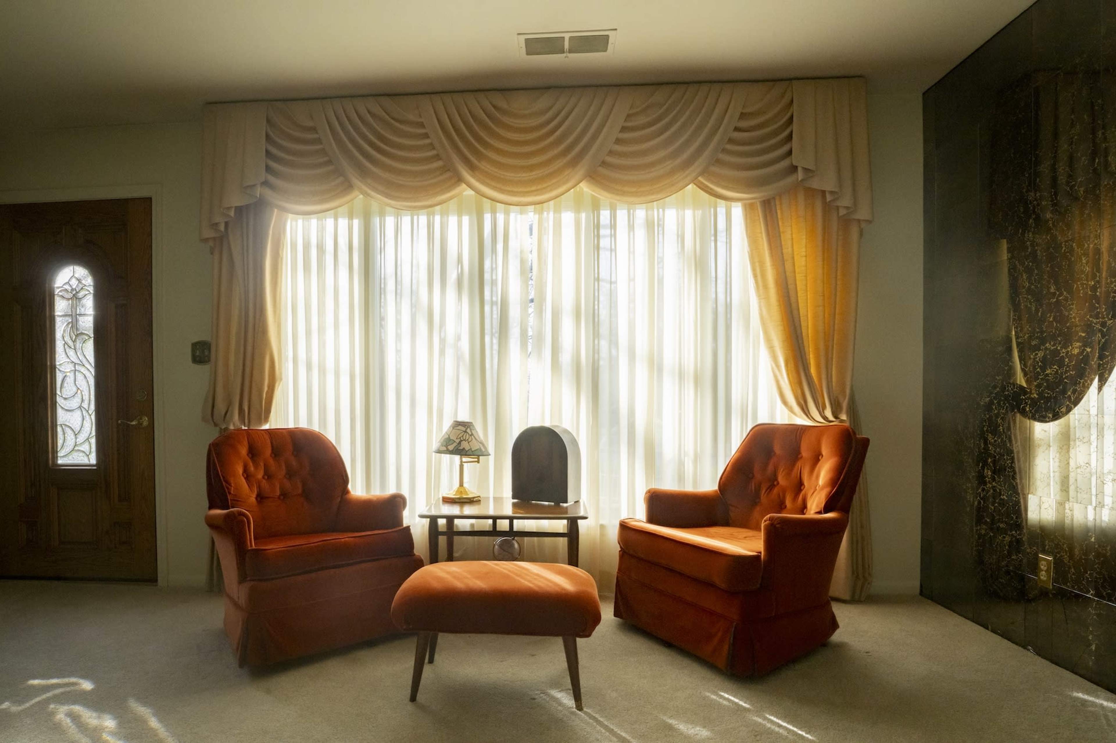 The image shows a living room with two orange chairs, a small table, and a lamp set in front of sheer curtains draped over a large window.