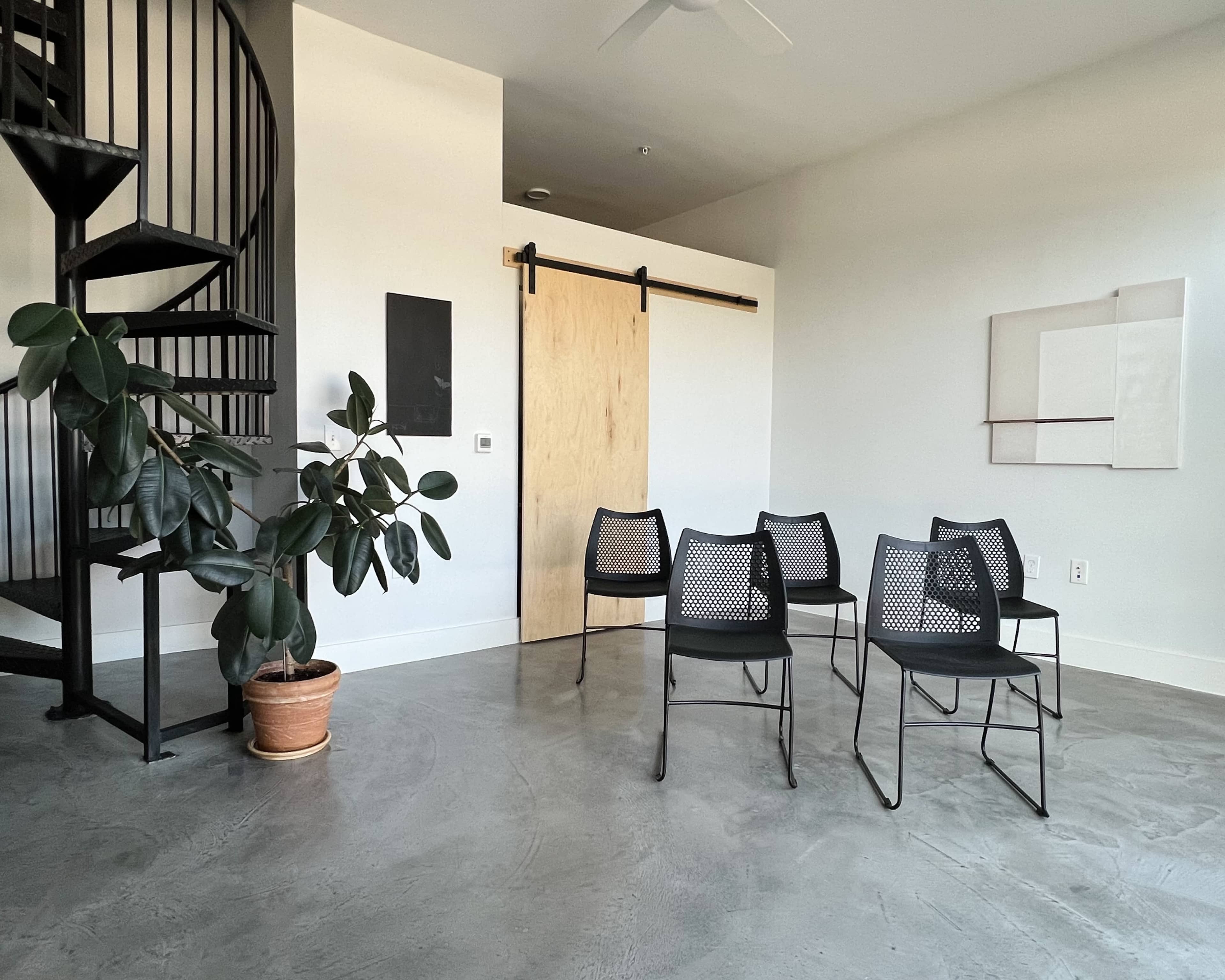 The image shows a modern, minimalist room with a spiral staircase, a potted plant, and six black chairs arranged around a central area.