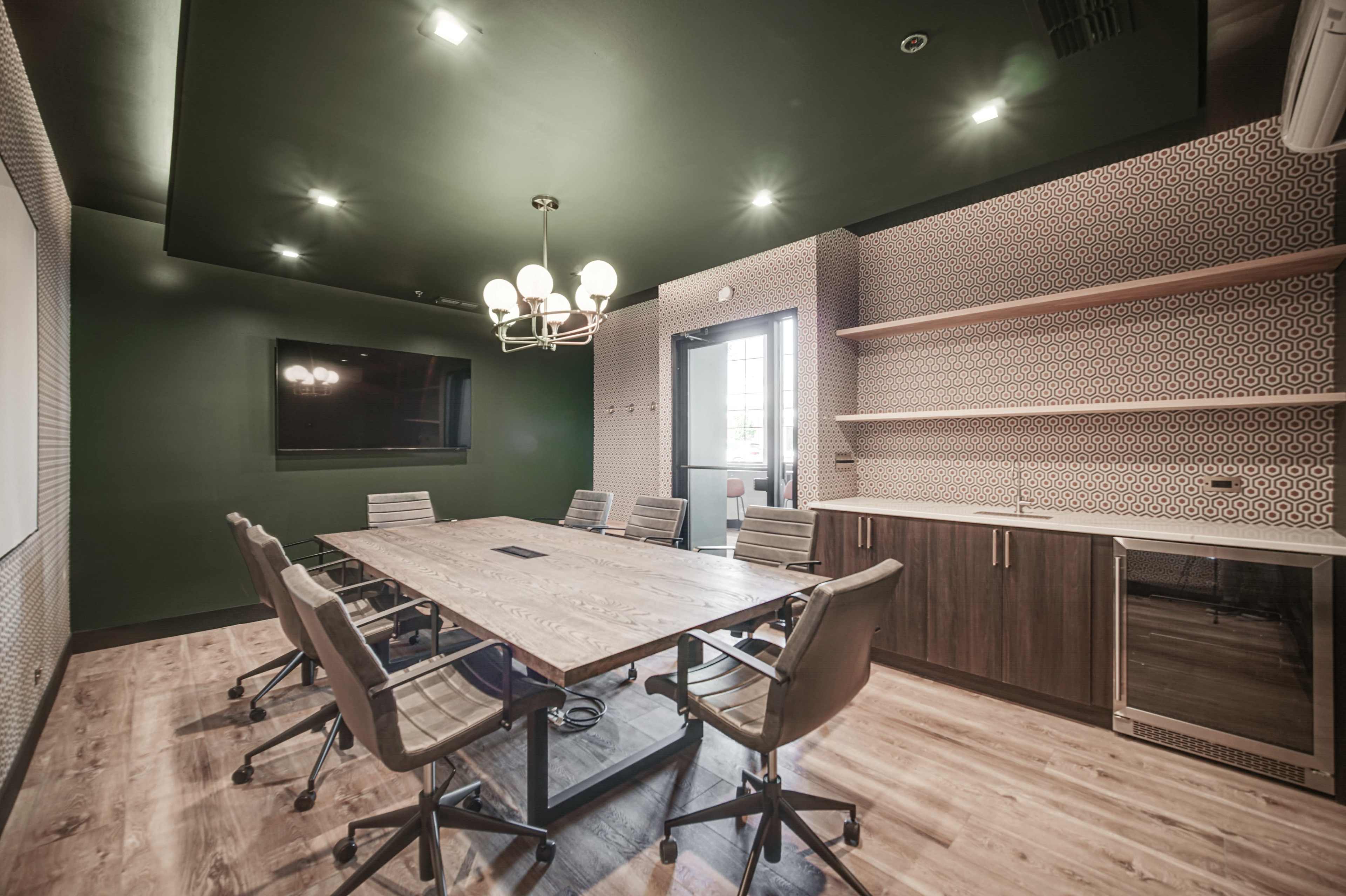 A modern conference room features a large wooden table surrounded by ergonomic chairs, with a wall-mounted screen and a kitchenette in the background.