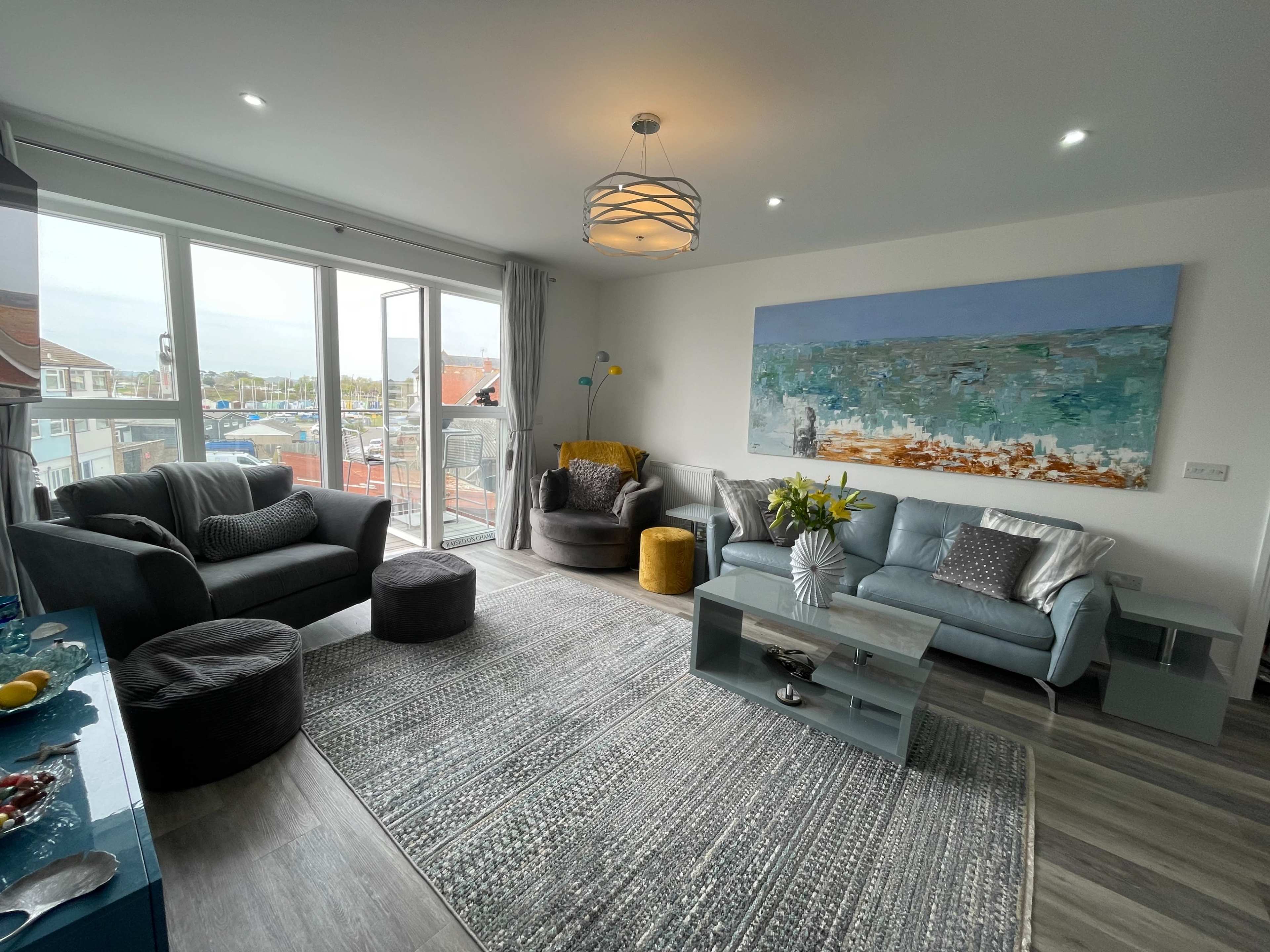 The living room features a teal sofa, two armchairs, a coffee table, and a large painting above the couch, with large windows overlooking an outdoor view.