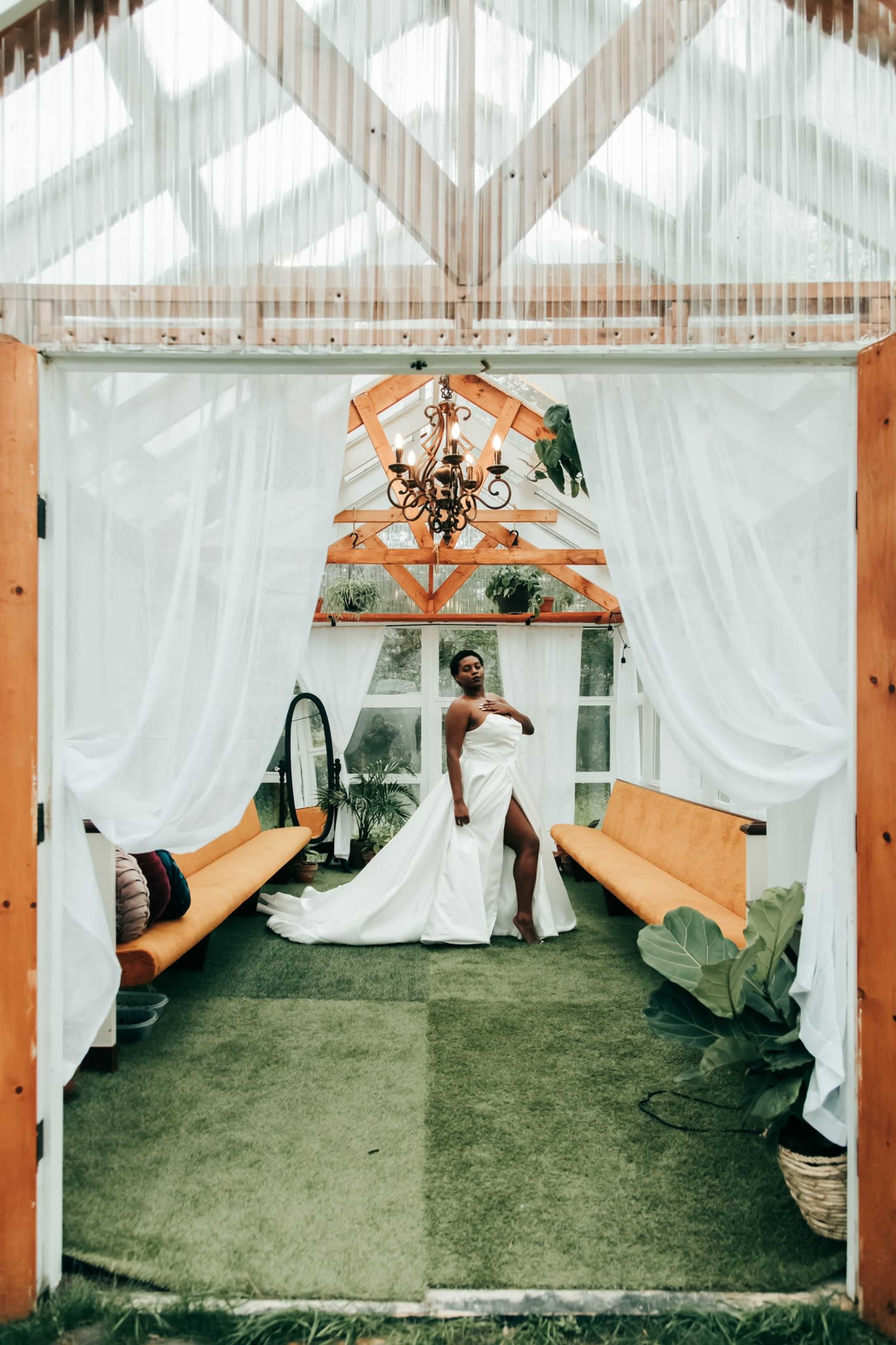 A bride in a white gown stands inside a greenhouse, with sheer curtains and wooden benches framing the scene.