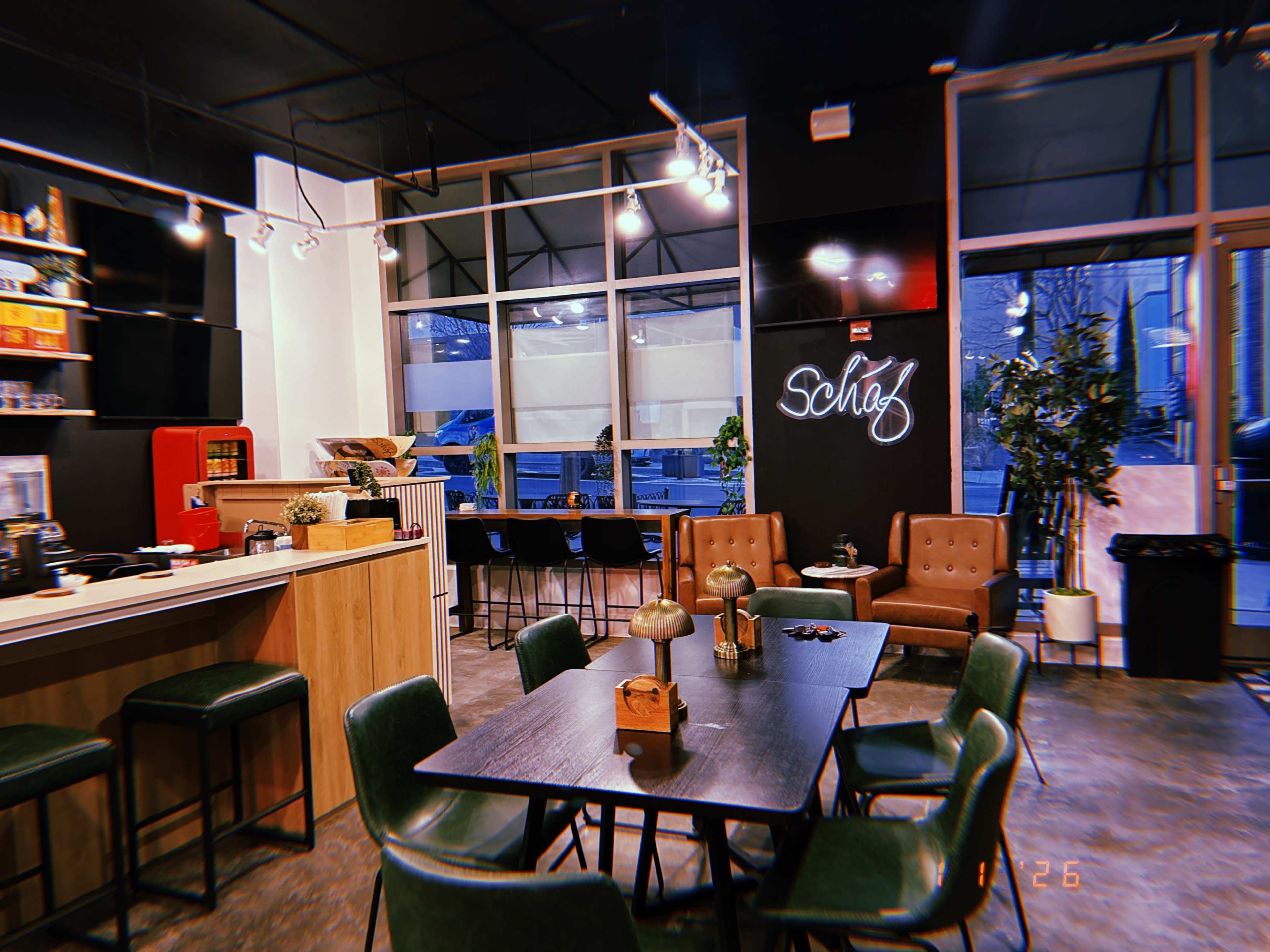 The image depicts a modern café interior featuring a coffee counter, several tables and chairs, and a neon sign that reads "Scholz."